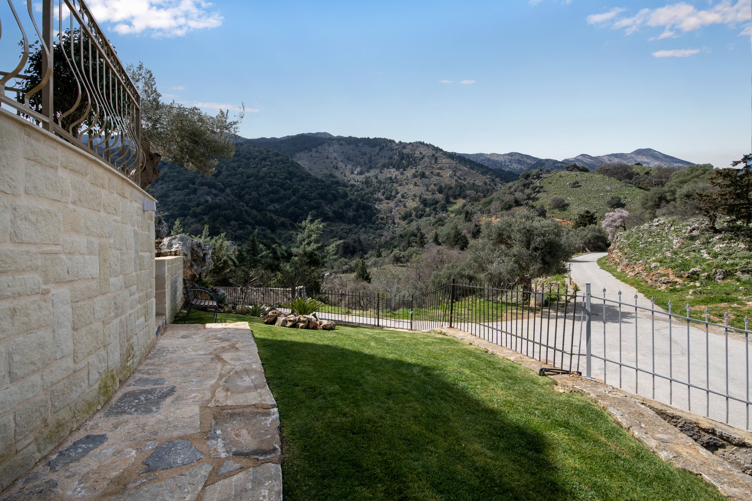 Stone patio with wrought iron railing overlooking lush green hills and mountains, sparse clouds in blue sky, gravel pathway on the right.