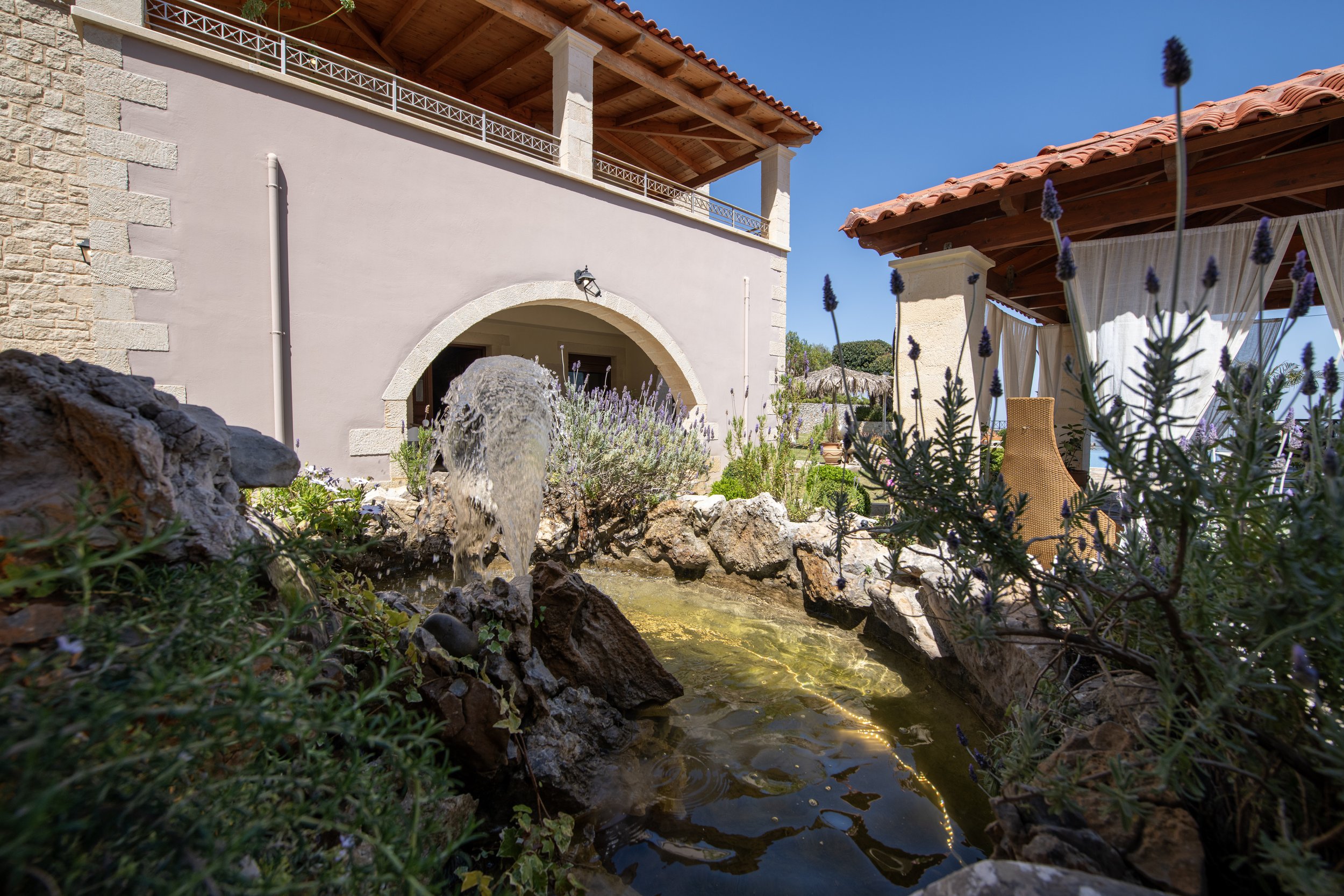 Outdoor patio with stone fountain, lavender plants, and beige building with arched entrance and wooden roof.