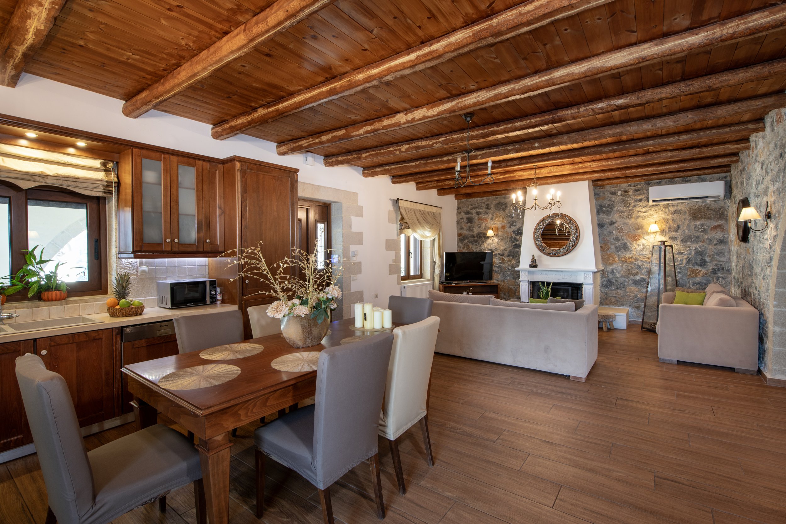 Rustic living room and kitchen with wooden ceiling beams, stone walls, and modern furniture, including a dining table, chairs, and sofa.
