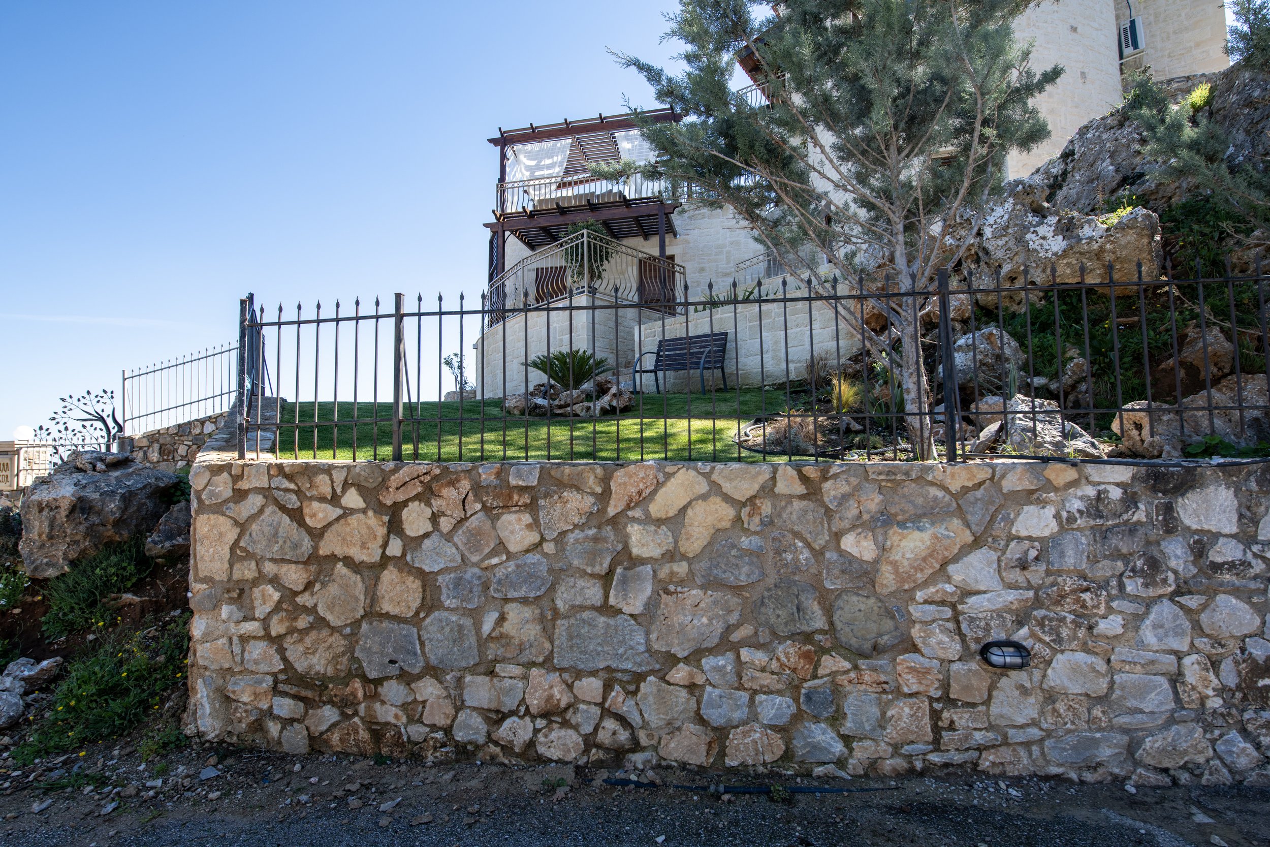 Stone wall with ornamental metal fence surrounding a landscaped area with lawn and trees, adjacent to a building with balconies.