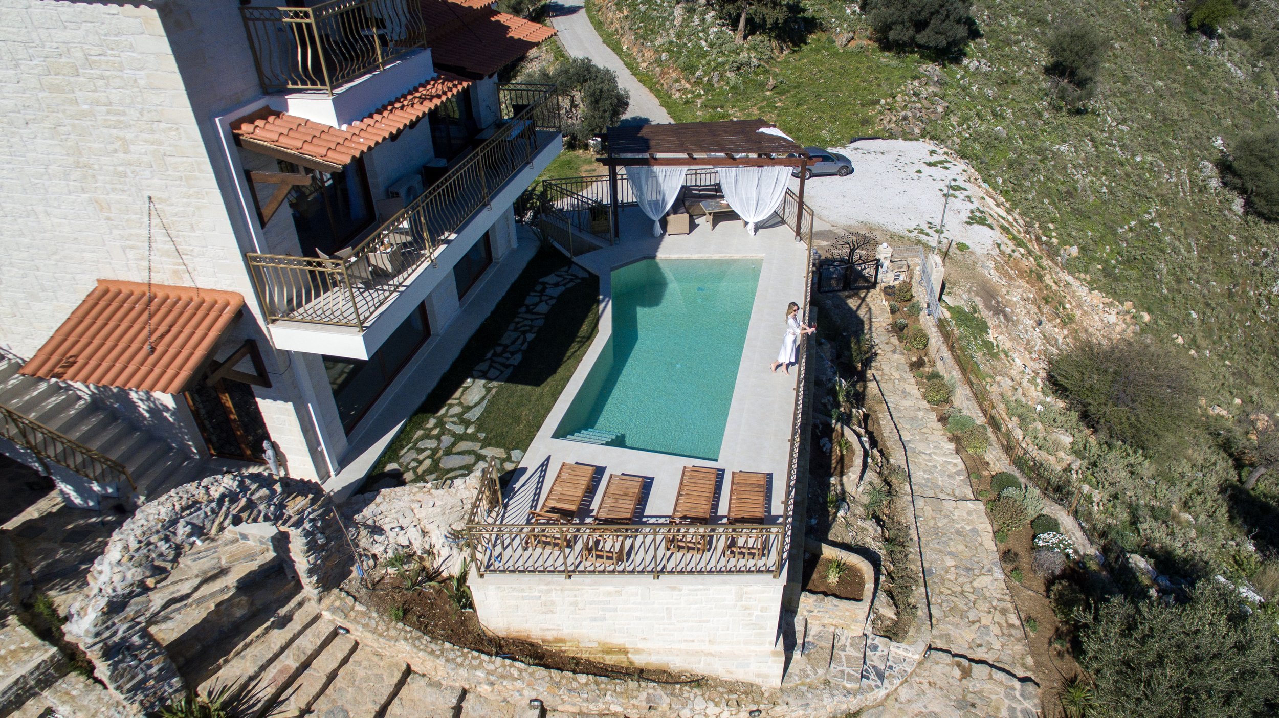 Aerial view of a luxury villa with a swimming pool, surrounded by a stone path and greenery on a hillside.