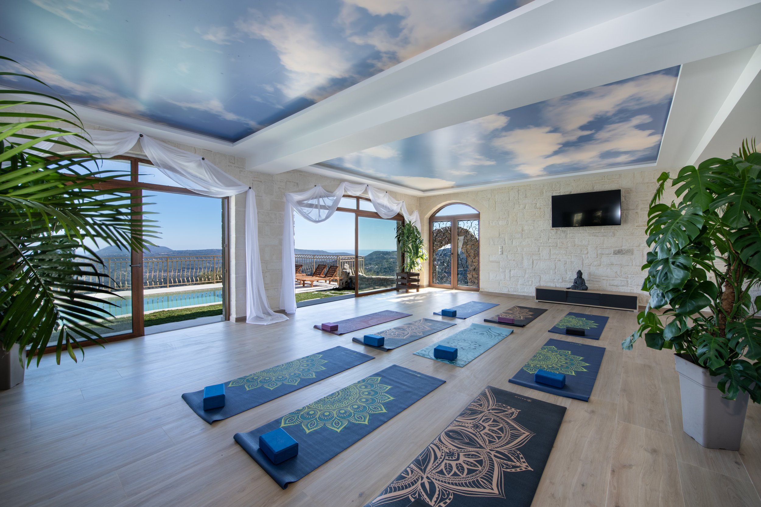 Yoga studio with decorative mats and blocks, a ceiling with a sky mural, large windows with a pool view, white curtains, and plant decor.