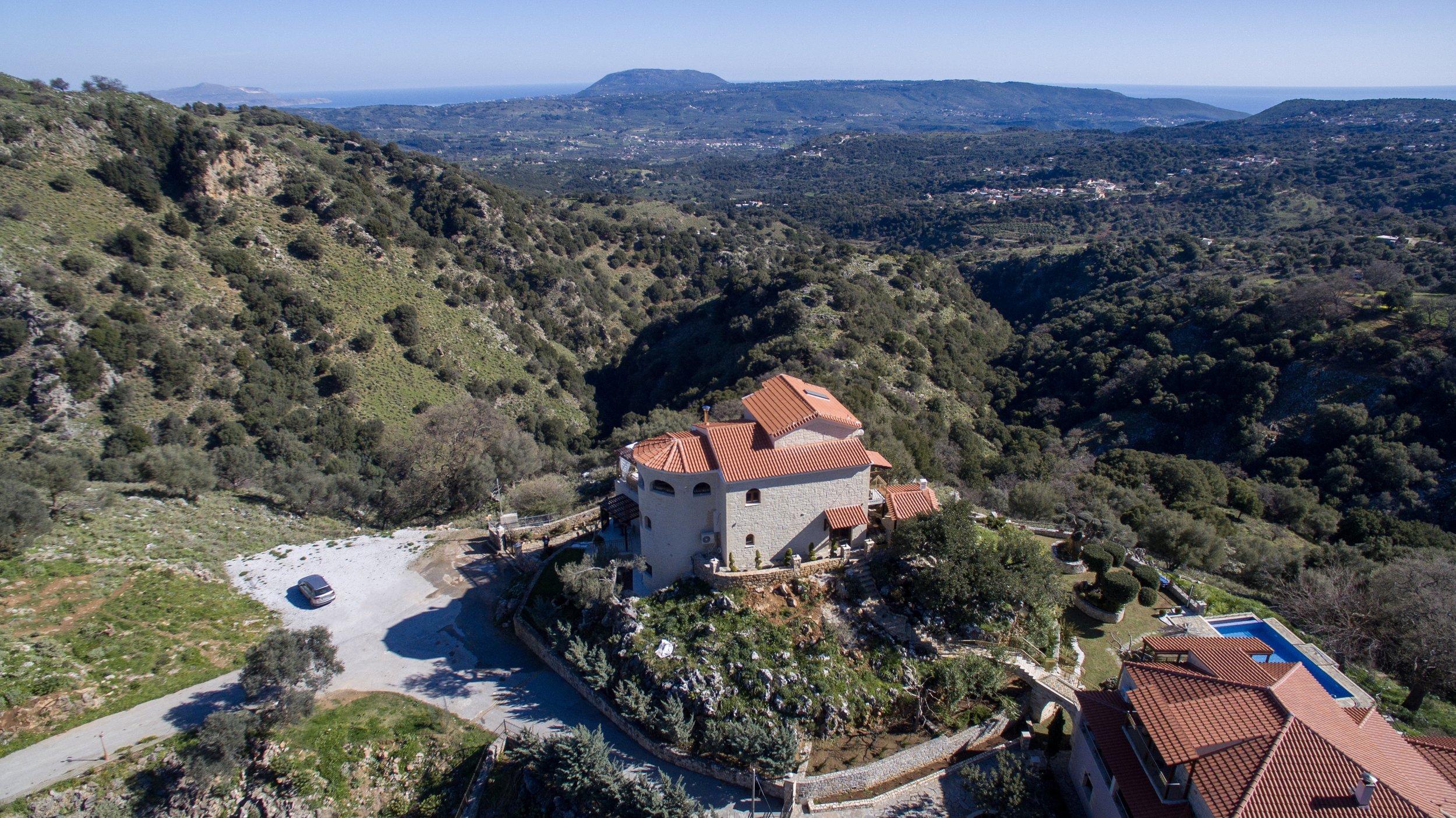 Aerial view of a large house with terracotta roof in a mountainous landscape with green vegetation and distant views of the sea.