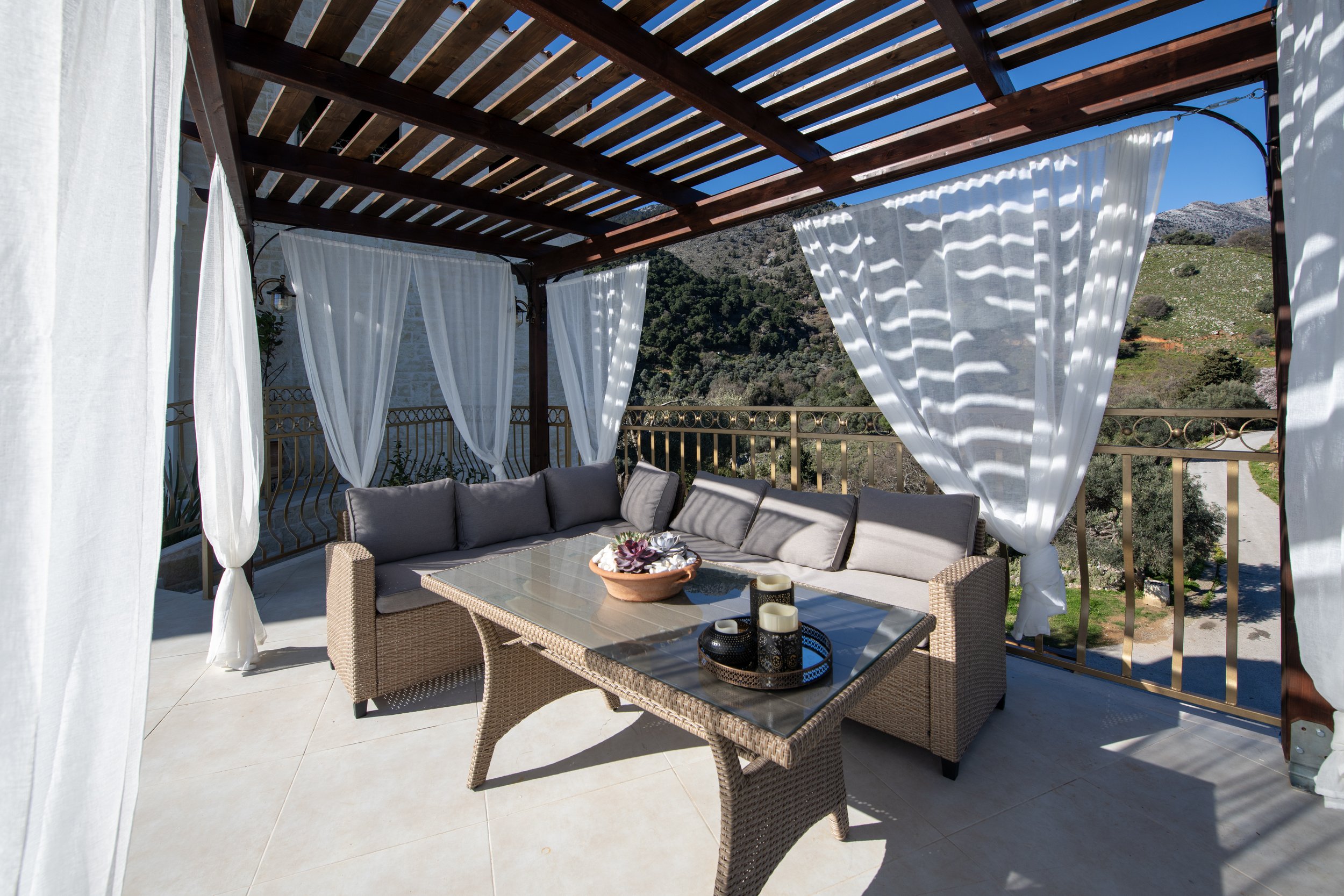 Outdoor patio with wicker furniture, glass-top table, and white drapes, under a wooden pergola with a mountain view.