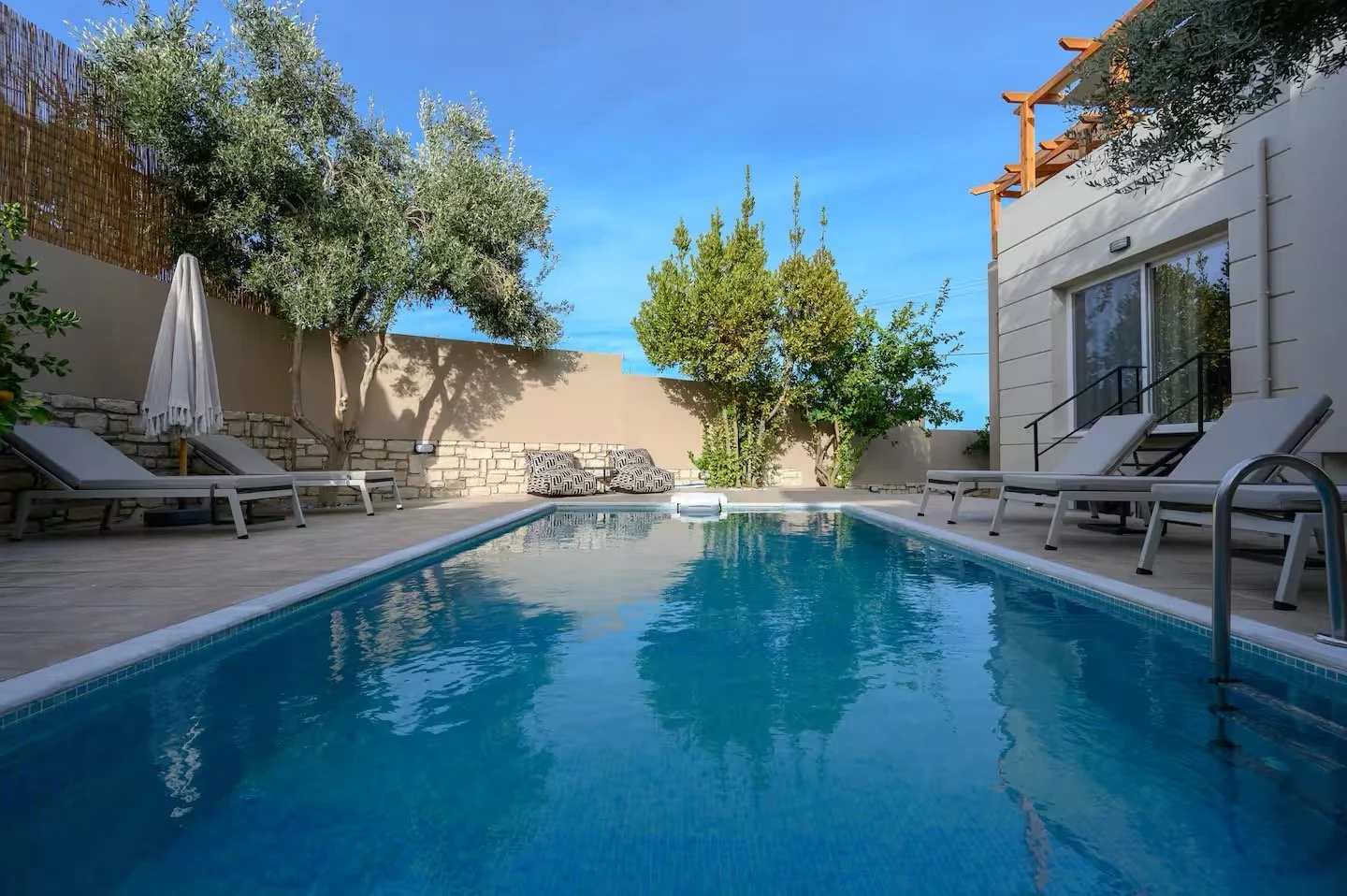Outdoor swimming pool surrounded by lounge chairs and umbrellas, with trees and a building in the background under a clear blue sky.