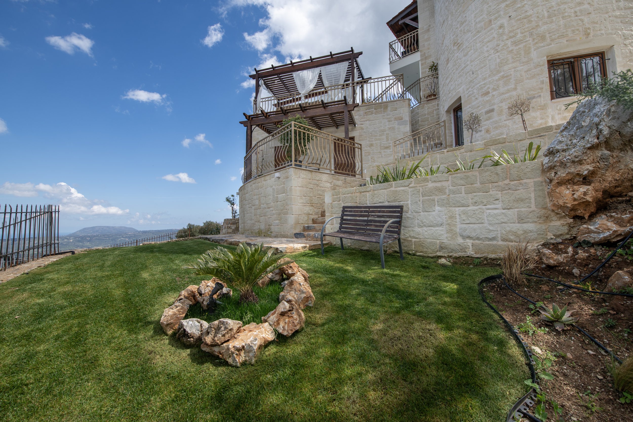 Stone villa with a pergola on a grassy hill, overlooking a scenic mountain view, featuring a wooden bench and rock garden.