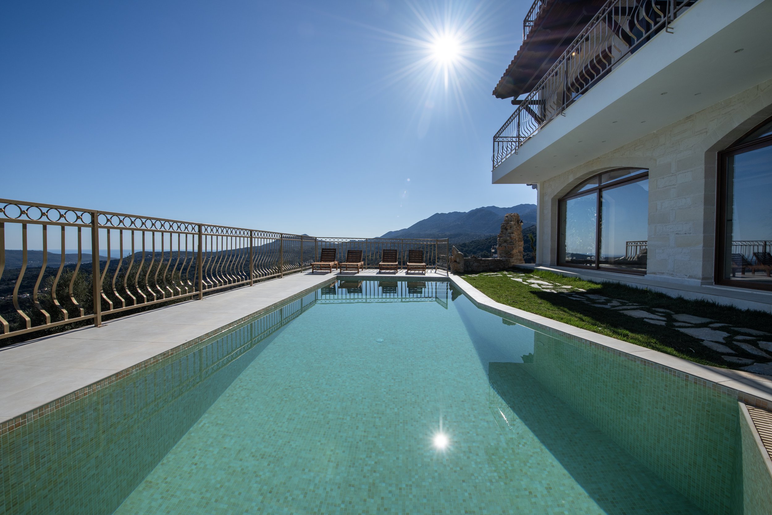 Infinity pool on a terrace with sunlit view of mountains and sky, surrounded by a modern building with large windows and deck chairs.
