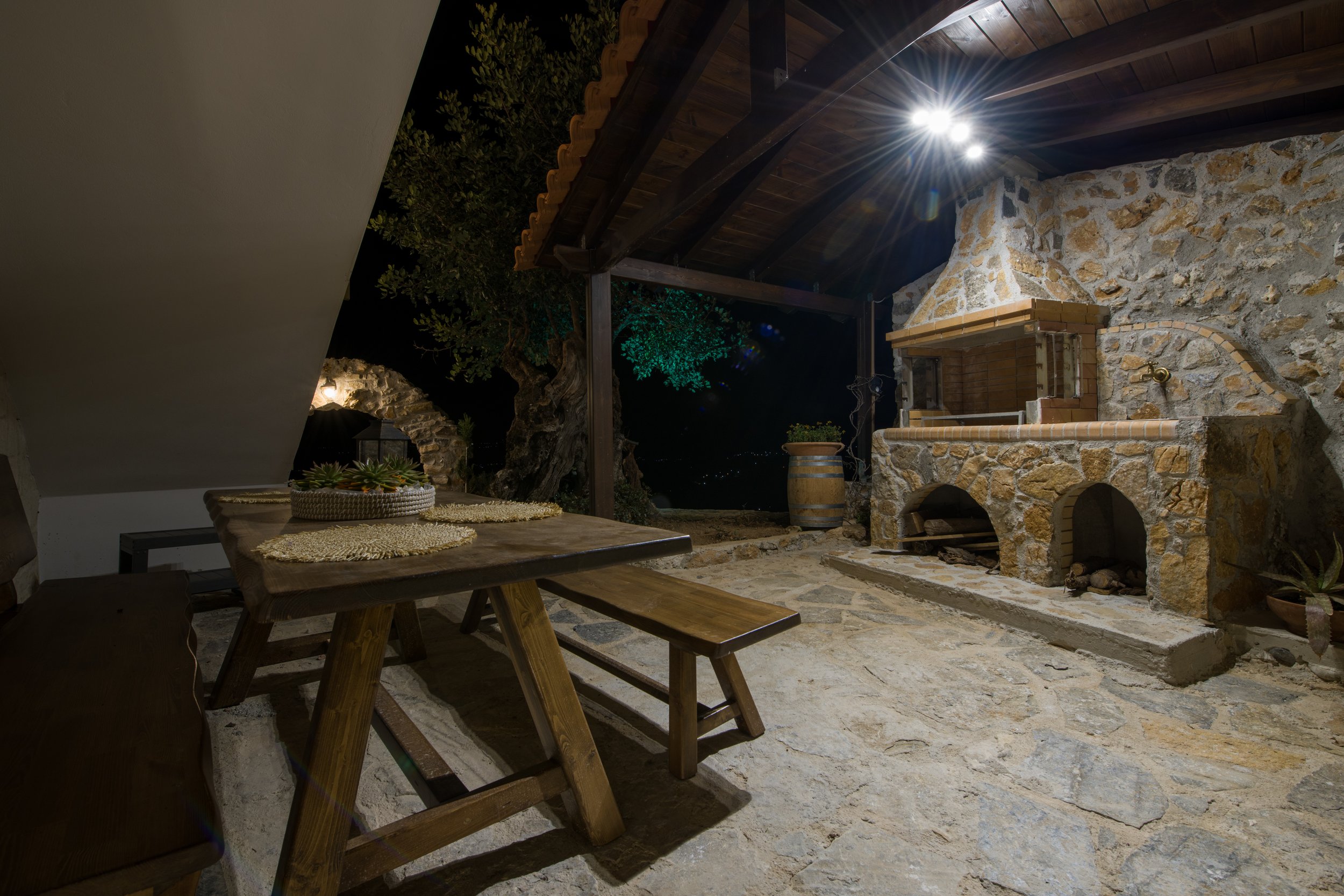 Outdoor stone patio at night with a wooden table and benches, stone fireplace, plants, and barrel under a wooden roof.