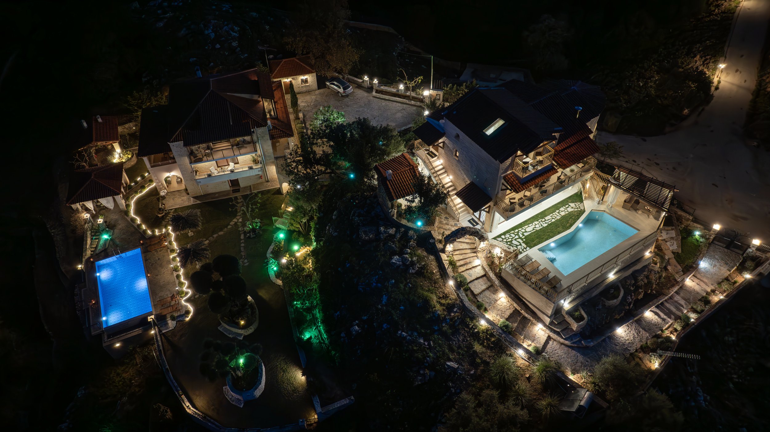Aerial view of two illuminated houses with swimming pools at night, surrounded by landscaped gardens and outdoor lighting.