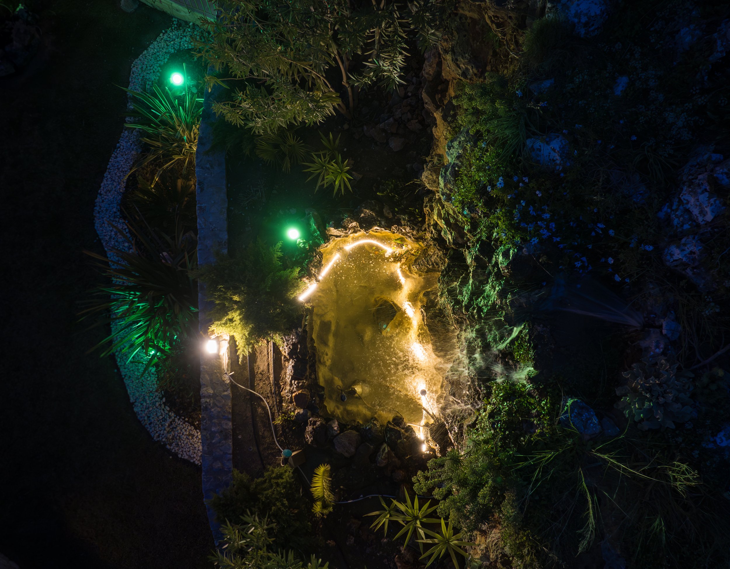 Aerial view of a garden pond illuminated by yellow and green lights at night. The pond is surrounded by rocks, plants, and a decorative pathway, creating a serene outdoor setting.