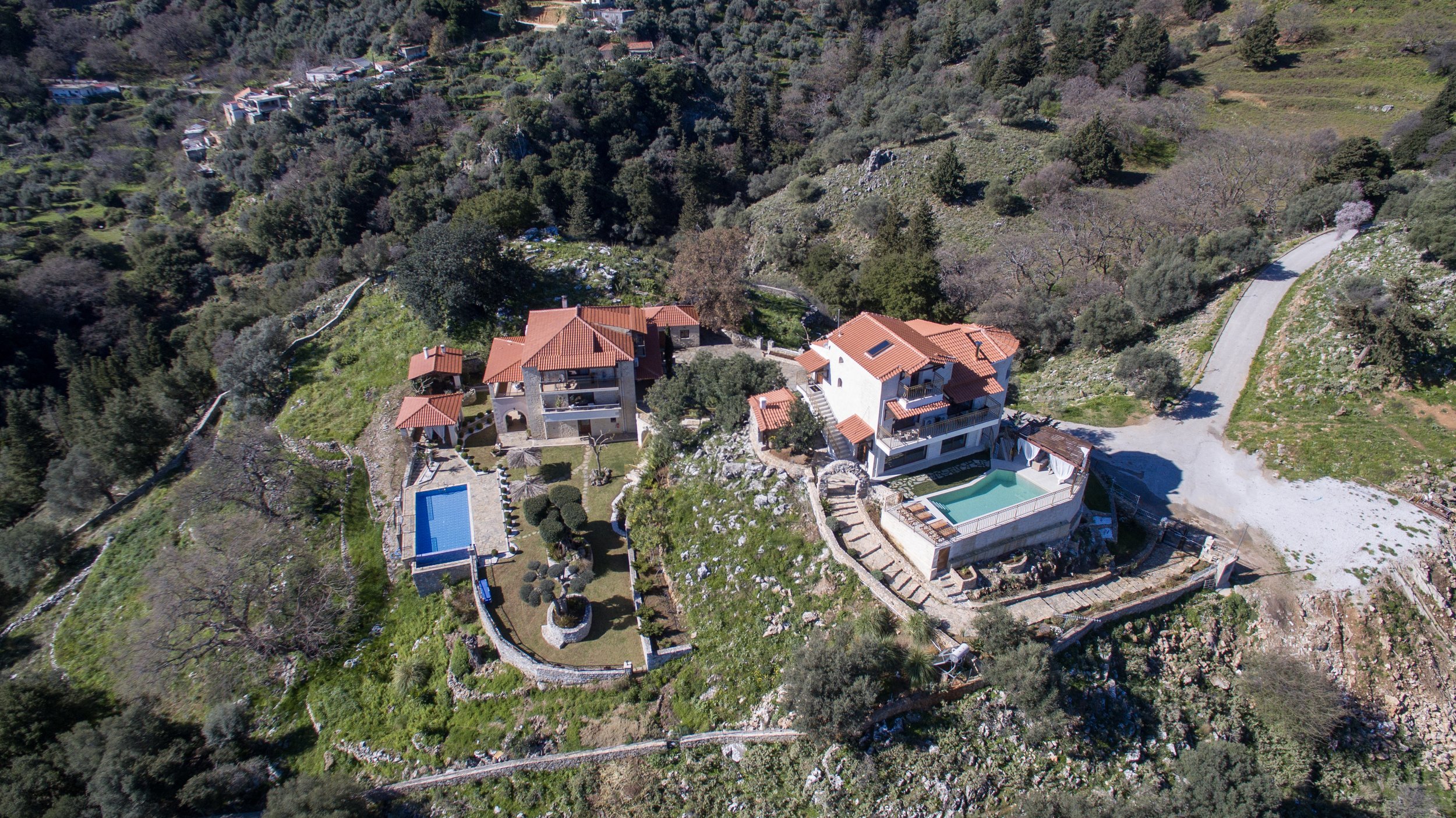 Aerial view of a hillside property with two buildings, swimming pools, red-tiled roofs, and surrounding greenery.