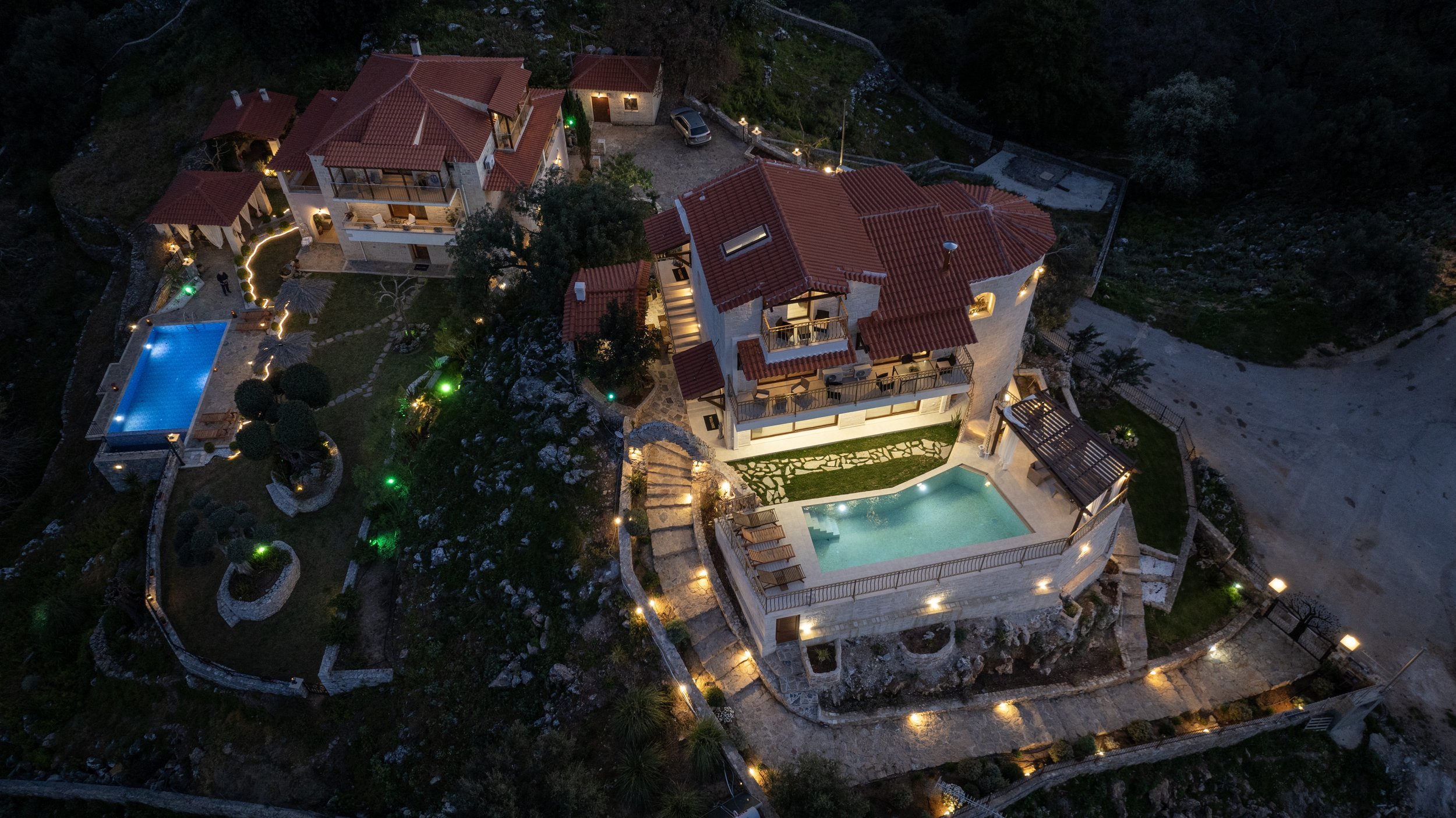 Aerial view of a luxury villa complex with swimming pools and landscaped pathways, illuminated by outdoor lighting at dusk. The villas have red-tiled roofs and multiple levels, set against a natural, rocky landscape.