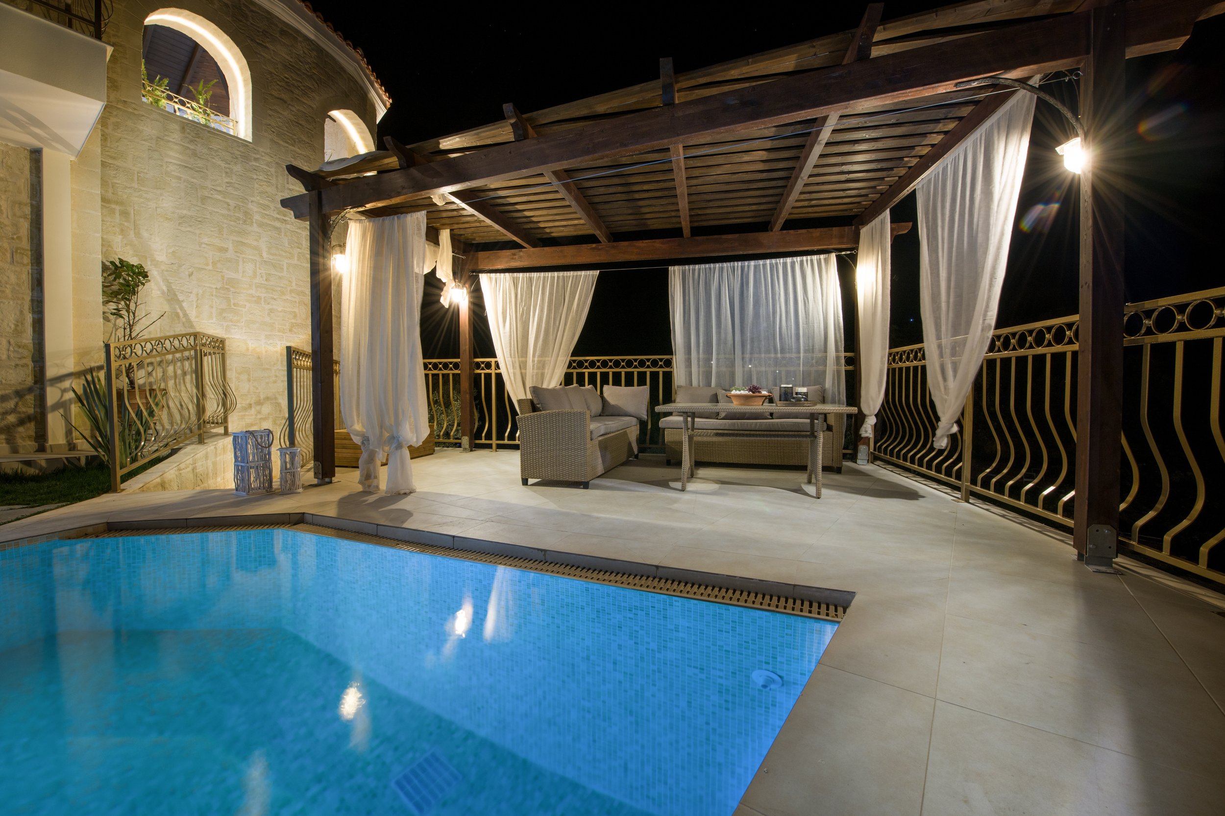 Outdoor patio at night with pool, wooden pergola, white curtains, rattan furniture, and stone house facade.