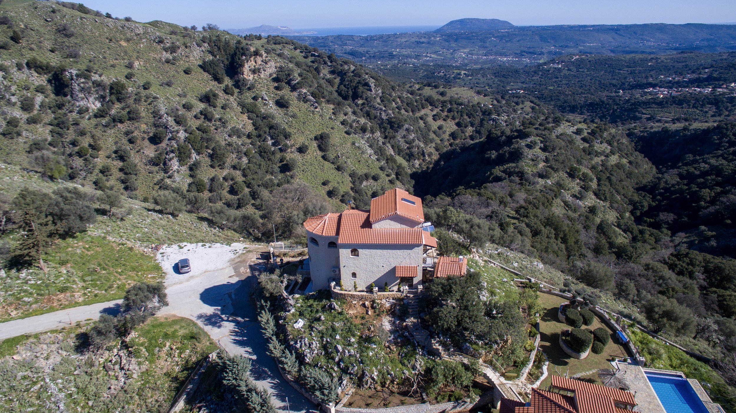 Aerial view of a house with a red tiled roof and stone walls located on a hillside. The surrounding landscape includes greenery, hills, and distant mountains. There is a swimming pool adjacent to the house, and a small parking area with a car nearby.