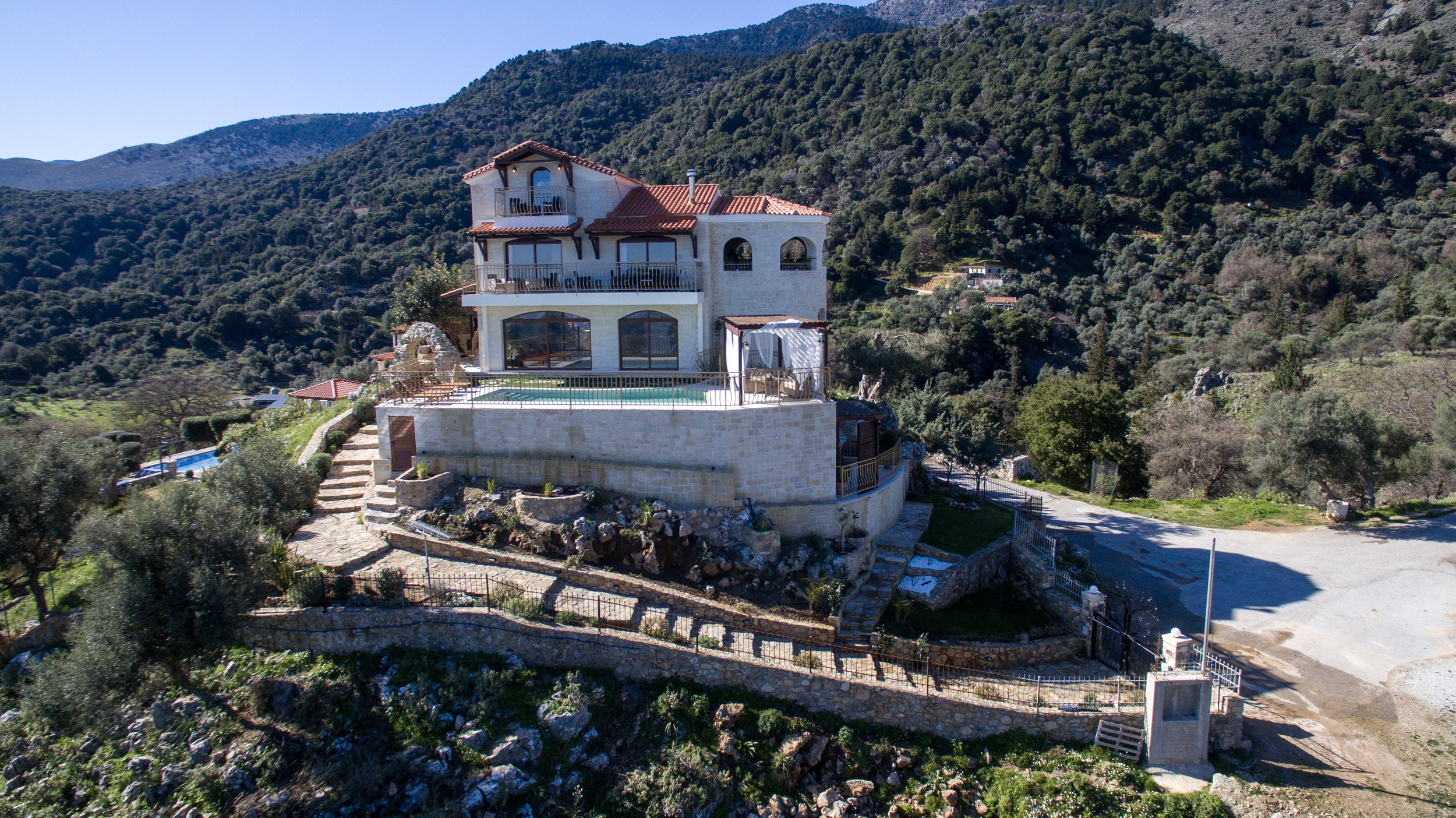 A large, multi-story house with a red roof and stone facade, situated on a hillside with a scenic view of mountains and forested landscape. The house features multiple balconies and a swimming pool on a terrace, surrounded by landscaped paths and gre