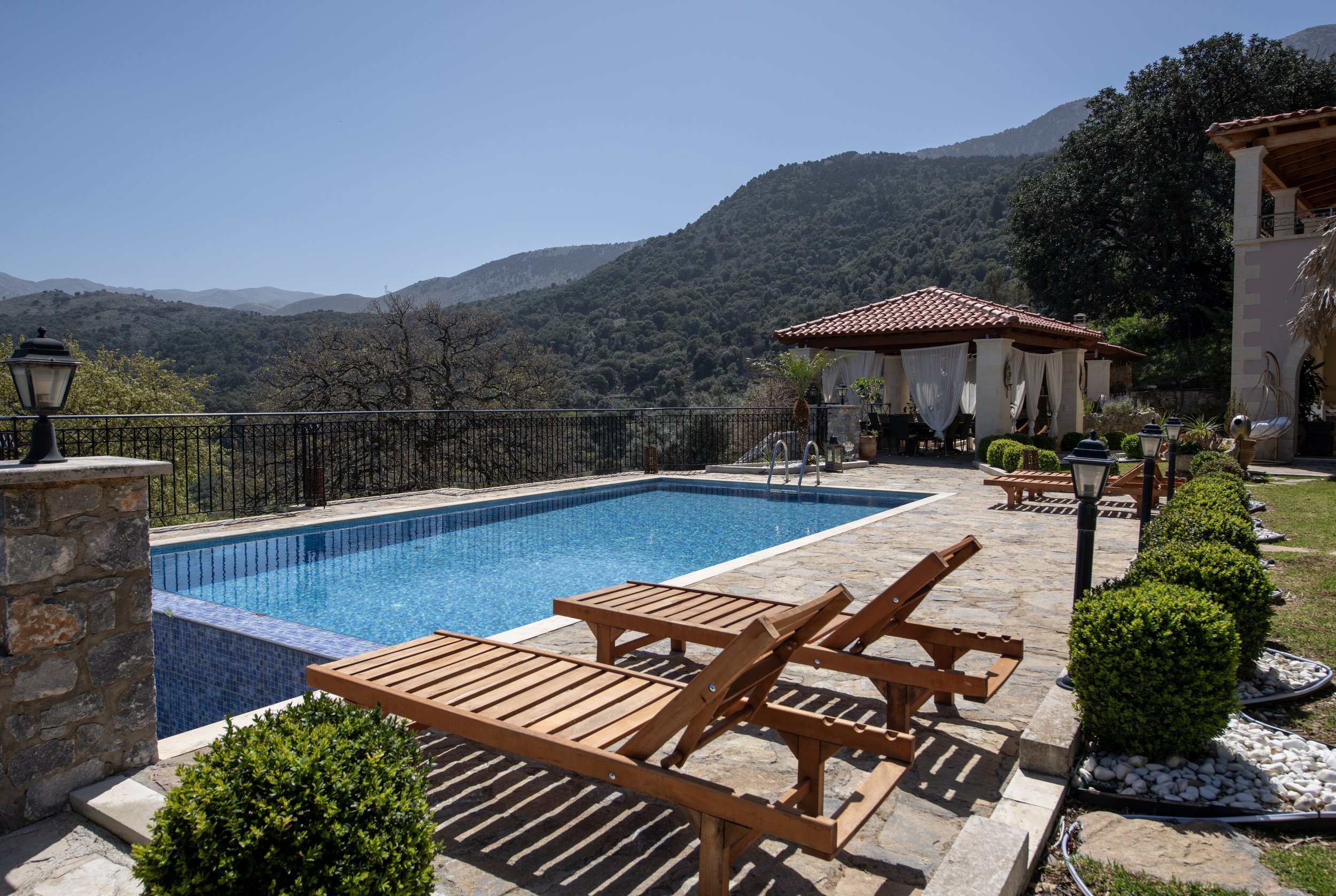 Outdoor swimming pool with wooden lounge chairs, surrounded by stone pavement, green bushes, and hillside landscape, with a gazebo in the background.