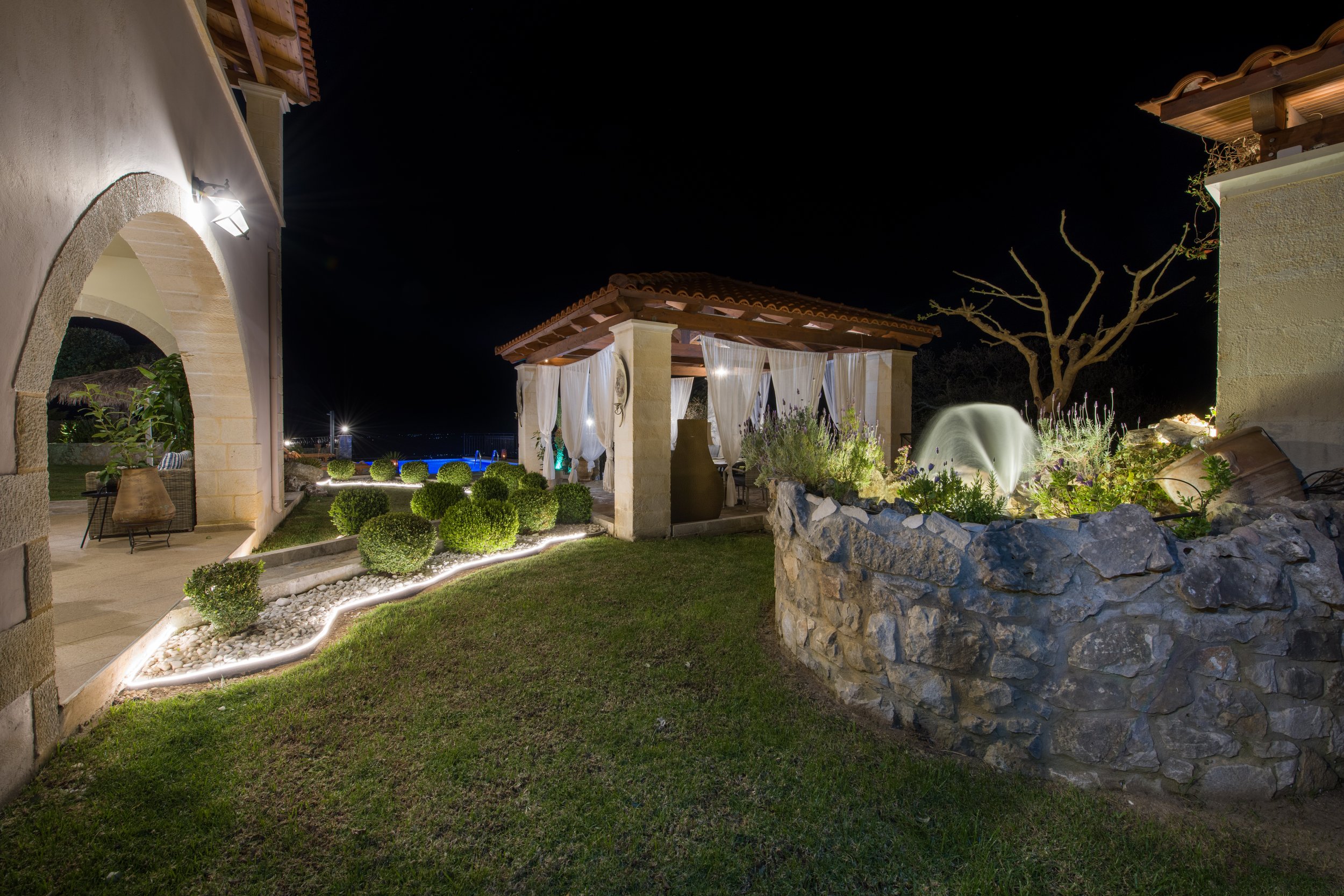 Night scene of a landscaped backyard with a stone patio, neatly trimmed shrubs, a pergola with white drapes, and ambient lighting.