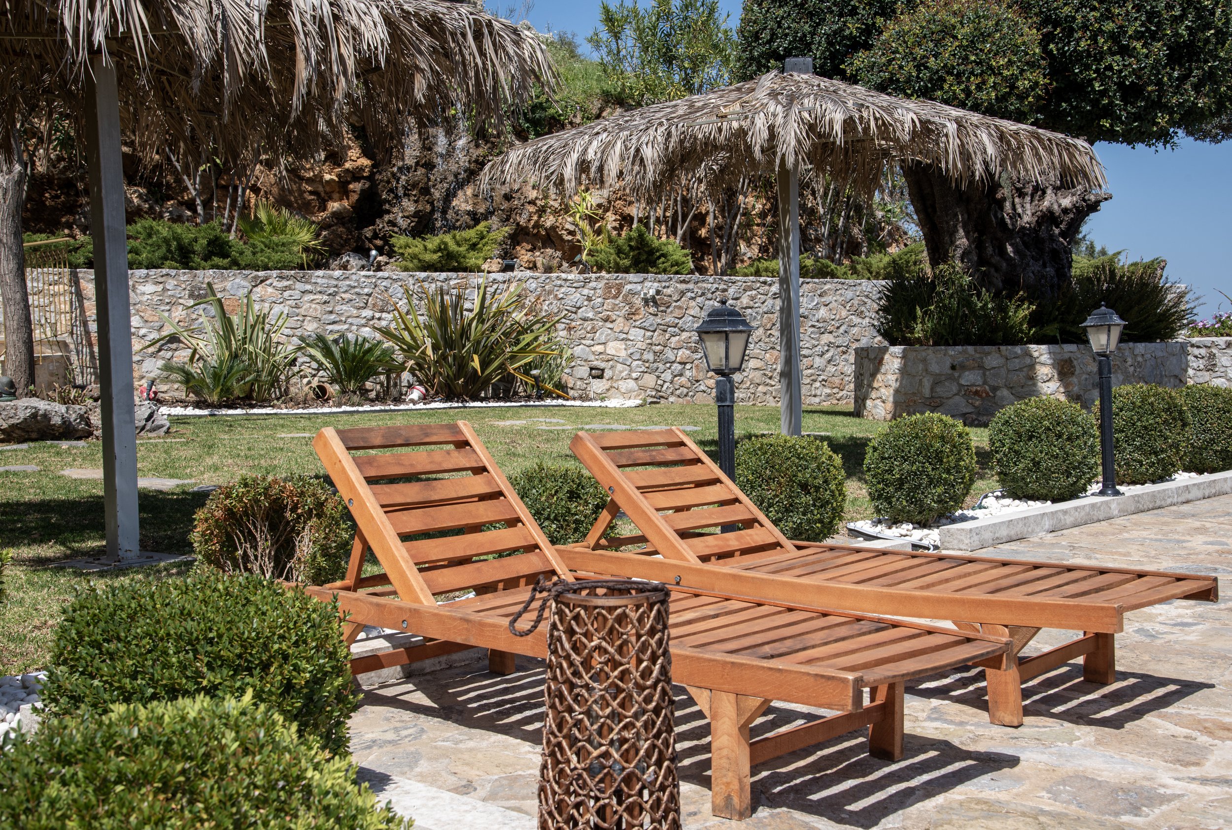 Wooden lounge chairs under thatched umbrellas on a stone patio in a landscaped garden with shrubs and stone wall.