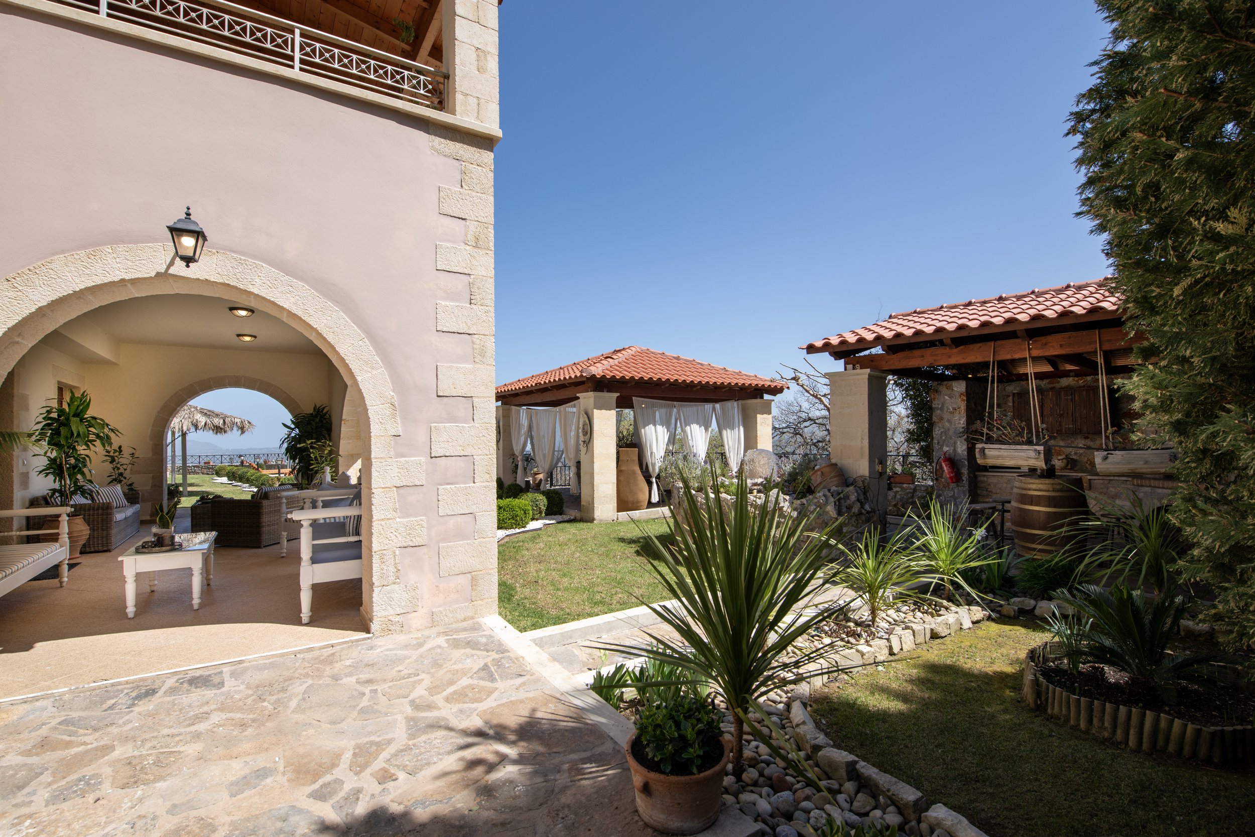 Outdoor patio with archway and seating area, manicured garden with potted plants, gazebo with curtains, and wooden pergola under clear blue sky.