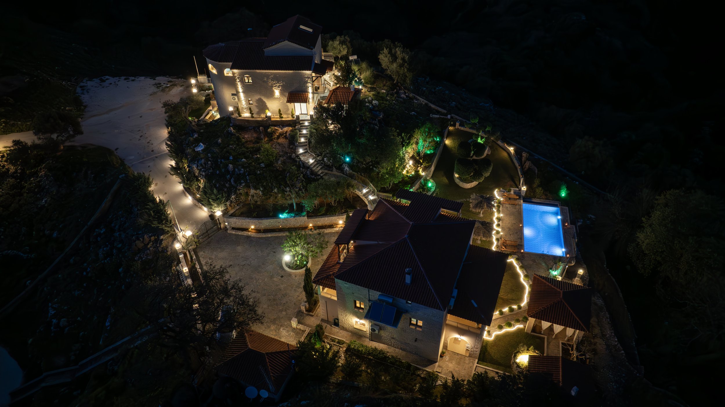 Aerial night view of an illuminated estate with buildings, pathways, a swimming pool, and landscaped gardens.