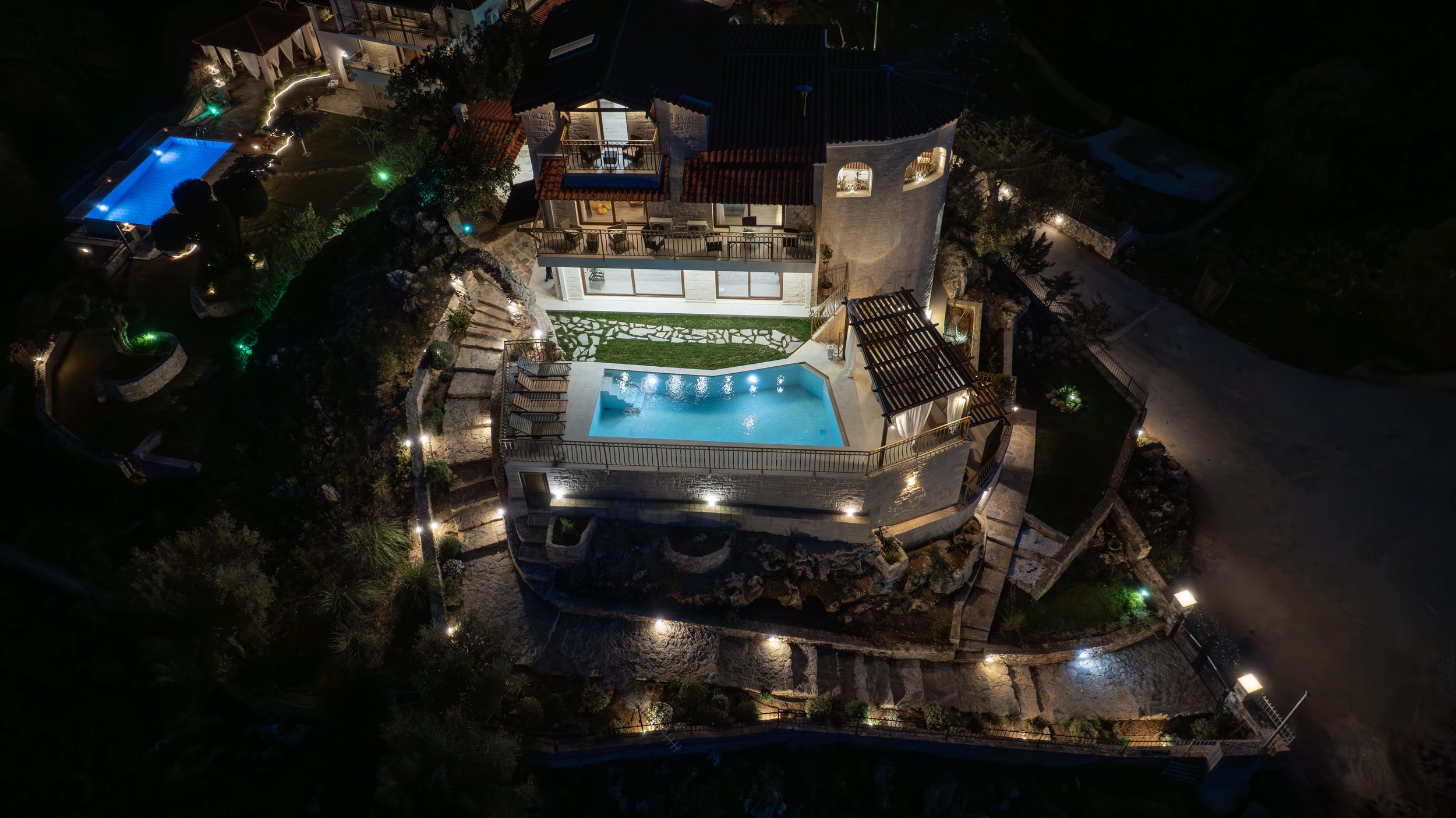 Aerial night view of a luxury villa with swimming pool illuminated, surrounded by landscaped gardens and outdoor lighting, situated on a rocky hillside.