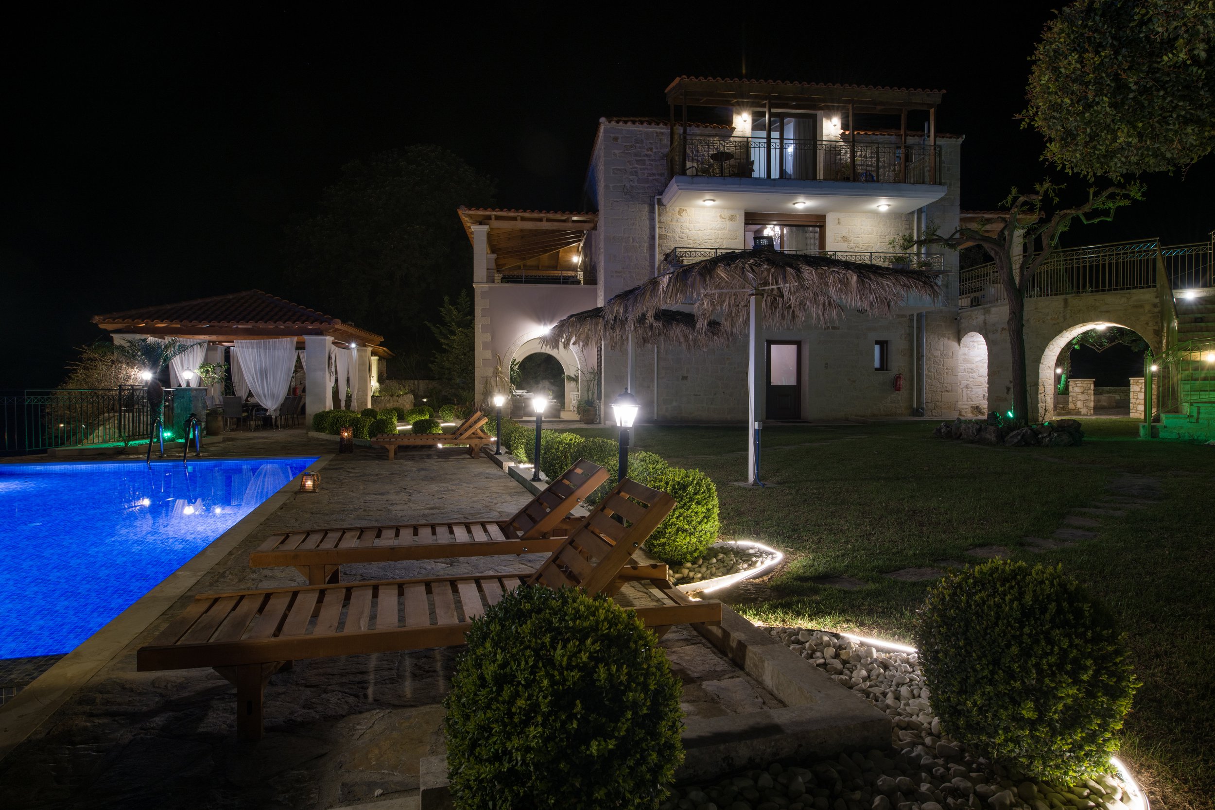 Night view of a luxurious villa with a lit swimming pool, wooden lounge chairs, and patio area with white curtains. The stone house features arched entrances and balconies, with well-manicured lawns and shrubs.