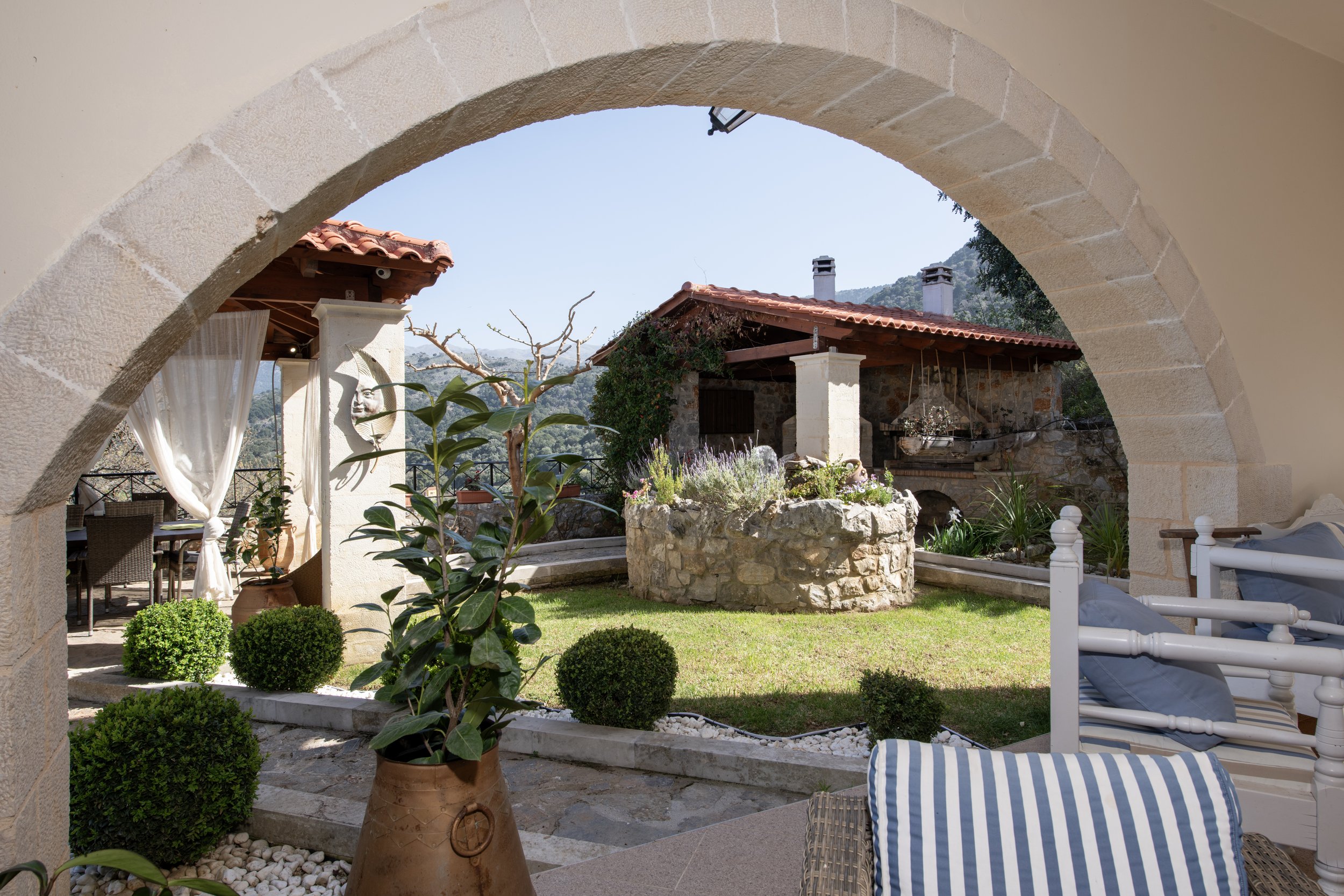 Stone archway leading to a garden patio with potted plants, stone fireplace, outdoor furniture, and mountain view.