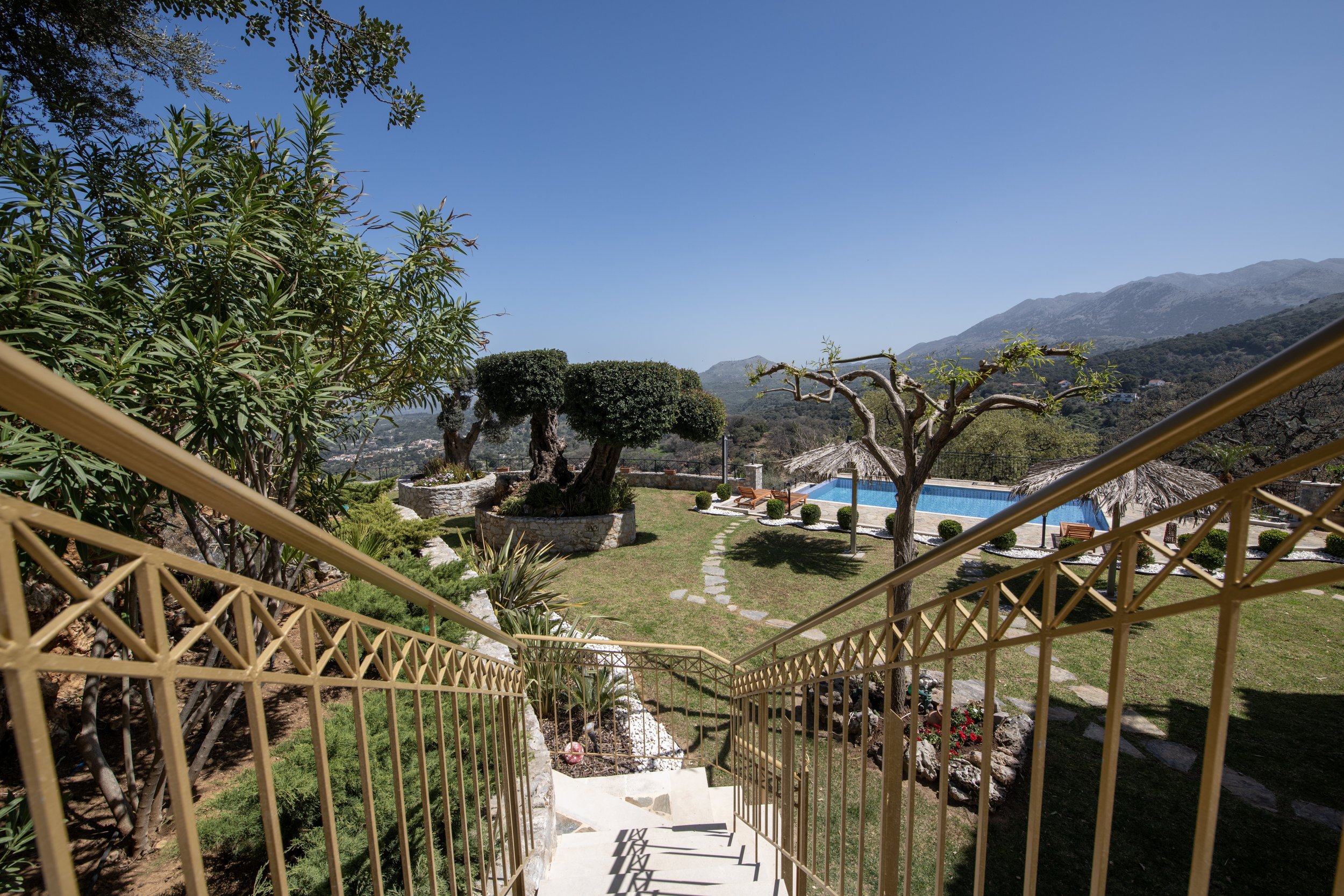 View from staircase with golden railing overlooking a landscaped garden with trees, a pathway, a swimming pool, straw umbrellas, and distant mountains under a clear blue sky.