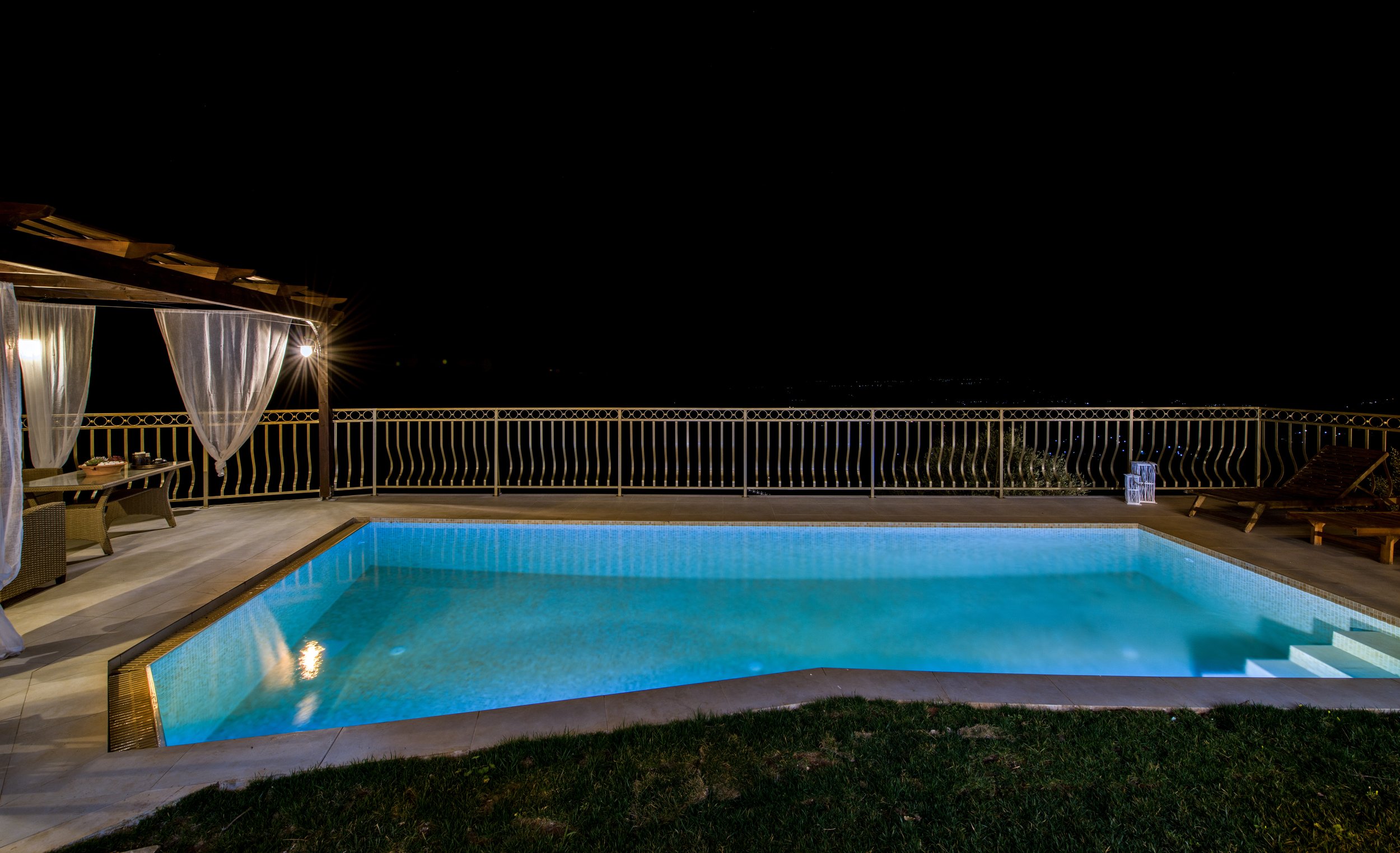 Nighttime view of an outdoor pool area with a lit swimming pool, wicker furniture, draped curtains, and a metal railing.