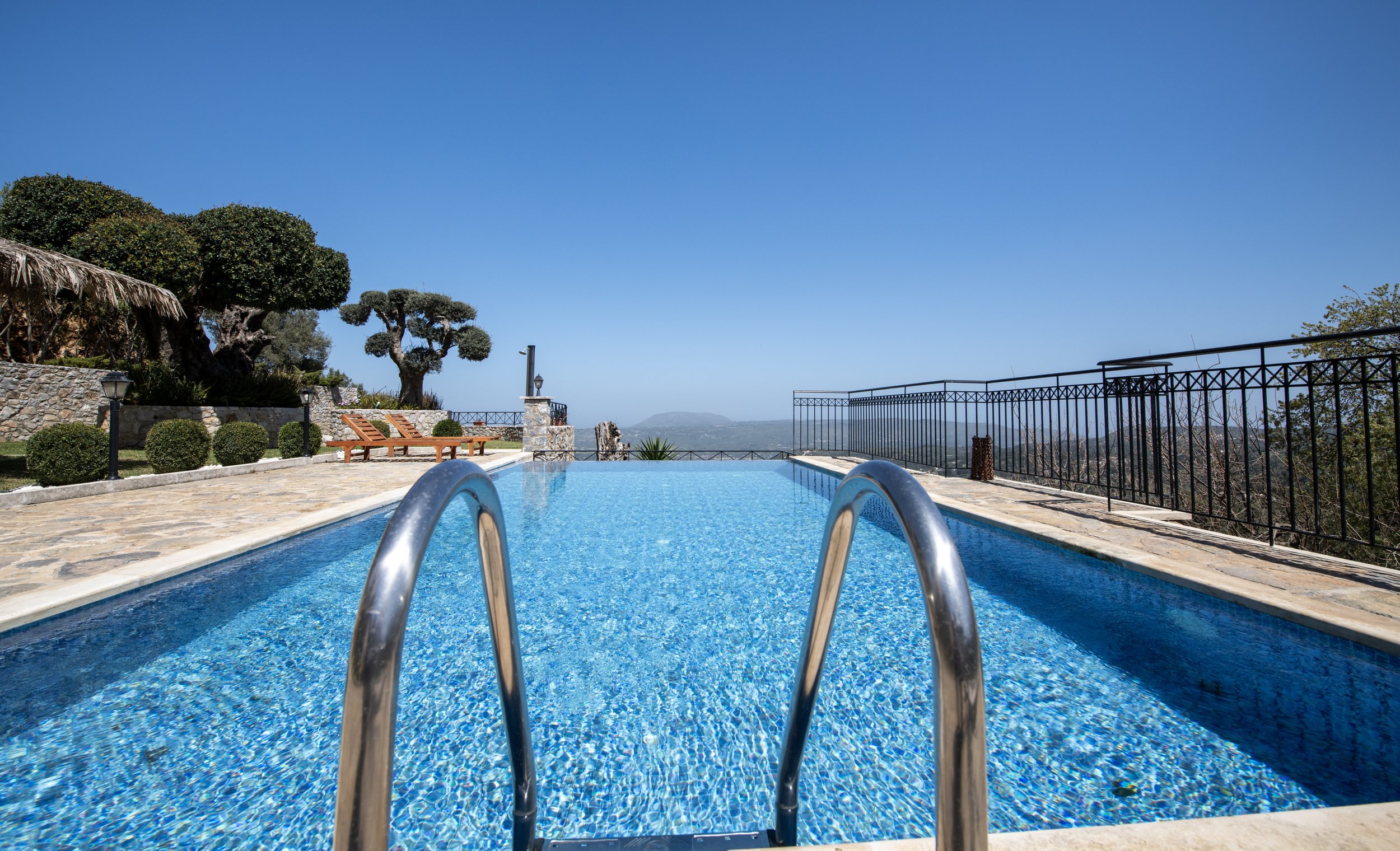 Luxury pool with metal ladder, overlooking scenic landscape and mountains, surrounded by stone patio and trees.