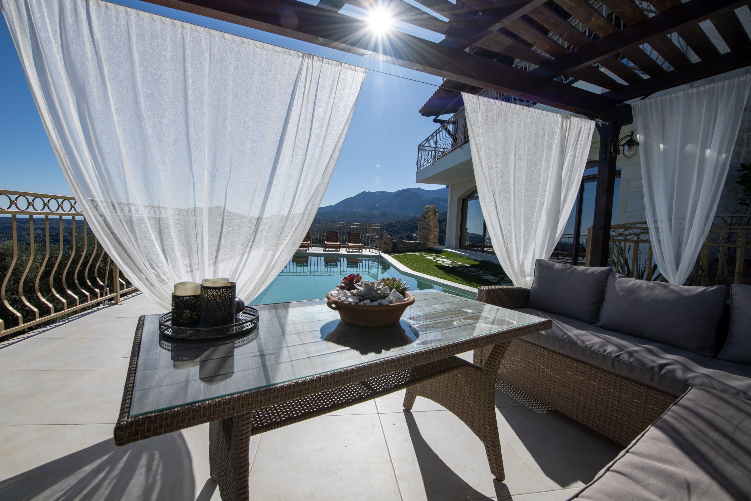 Luxury patio with glass table, white curtains, and wicker furniture overlooking a pool and mountains under a bright sun.