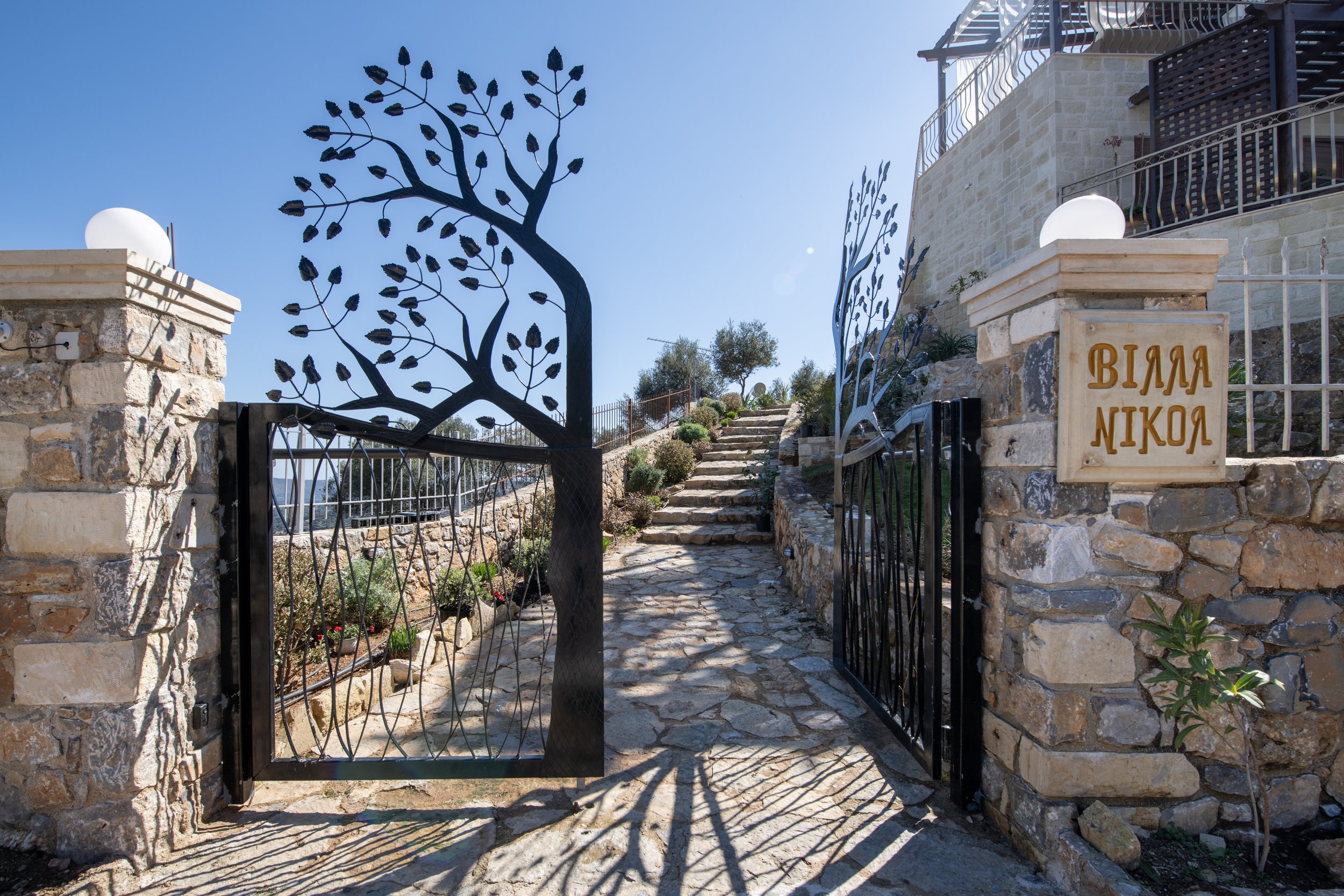 Decorative metal gates with a tree design open to reveal a stone path leading up to a house, with "Villa Nikola" written on a stone pillar.