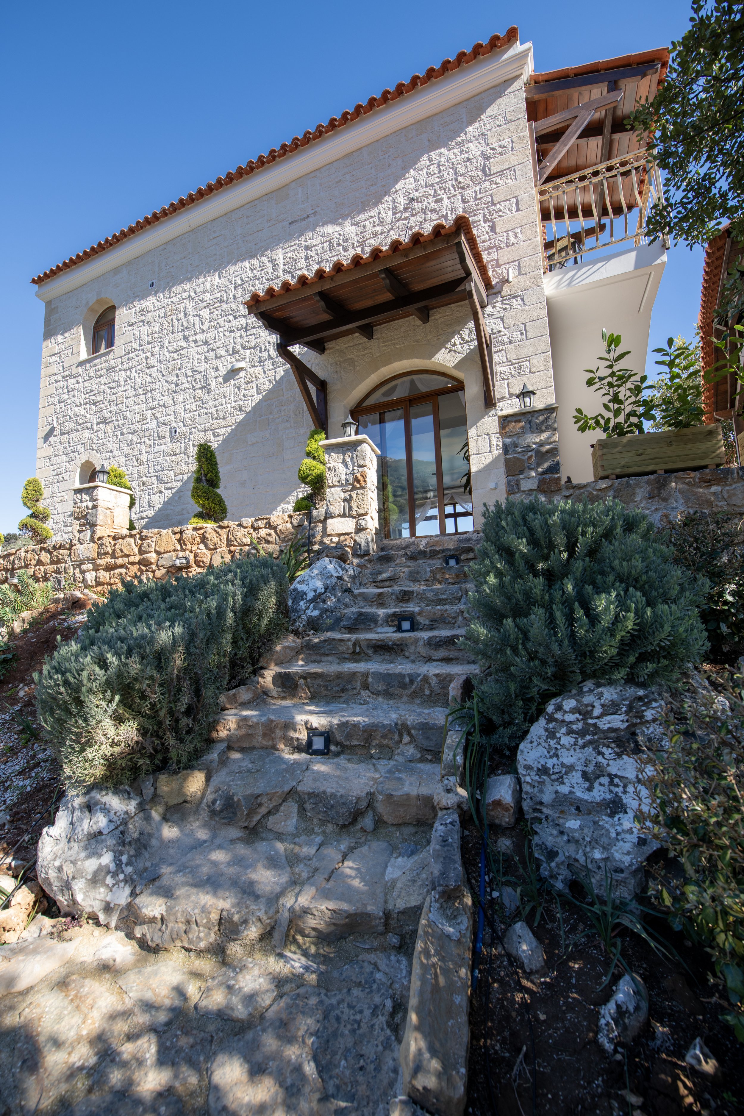 Stone staircase leading to a Mediterranean-style house with a tiled roof and arched wooden door, surrounded by green shrubs and trees, under a clear blue sky.