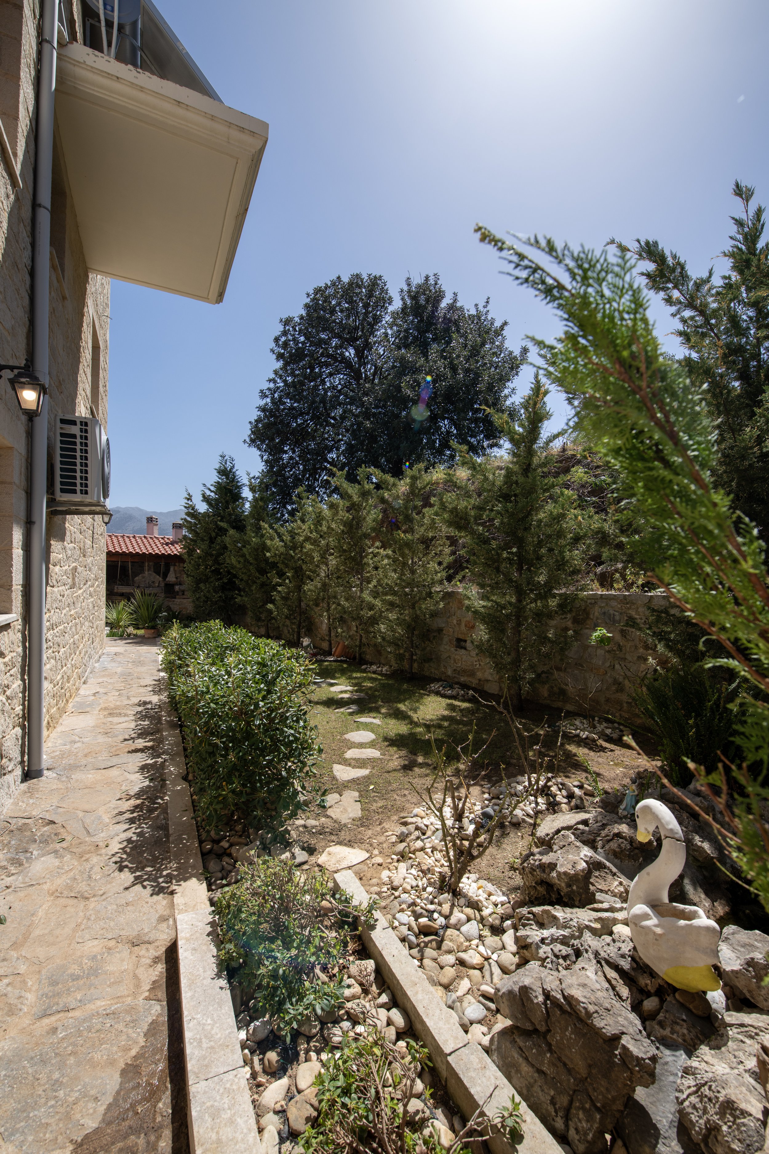 Side view of a landscaped garden with stone pathway, decorative swan, and green shrubs next to a stone building under a sunny sky.