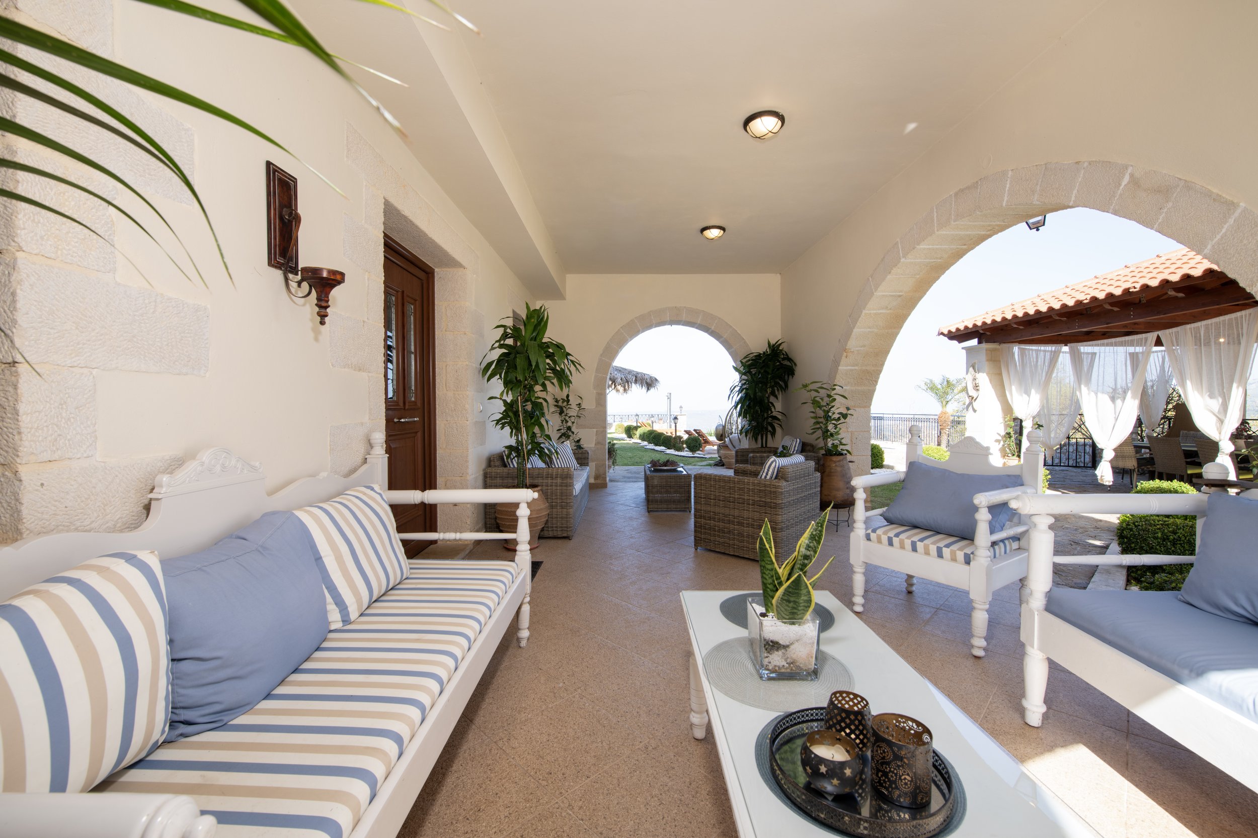 Outdoor patio with striped cushions, white sofas, plants, and decorative items, featuring arches and a view of a garden.