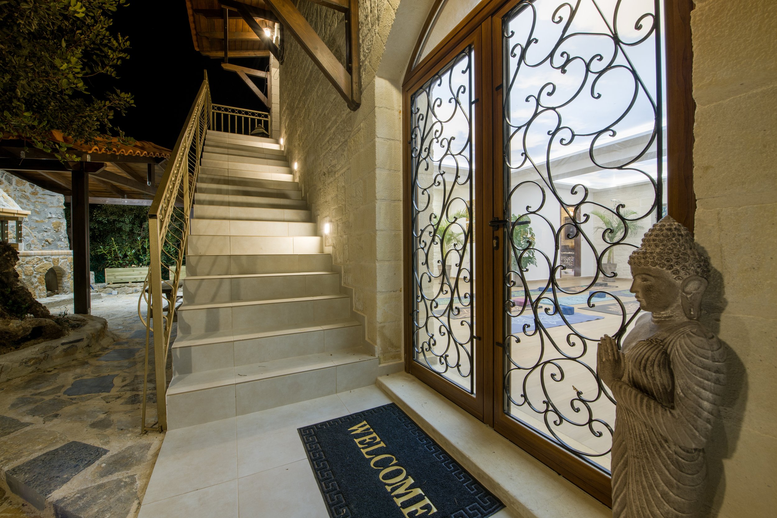 Indoor stairway with a welcome mat and a decorative metal grille door, featuring a statue of Buddha. Exterior view includes stonework and greenery.