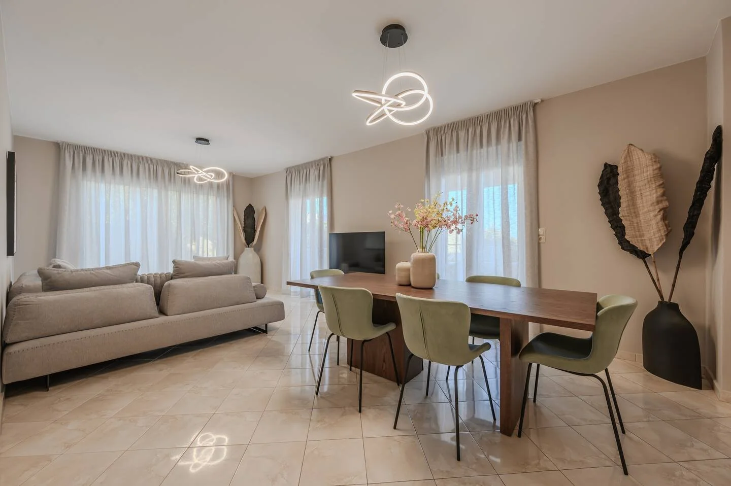 Living room with a beige sofa, a wooden dining table with six green chairs, a flat-screen TV, modern ceiling lights, and decorative items like large vases and wall art, with light-colored curtains and tiled floor.
