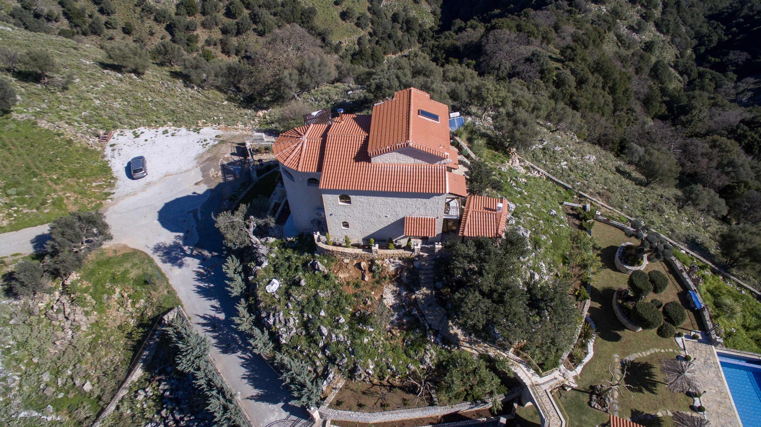 Aerial view of a large stone house with terracotta roof tiles surrounded by greenery and mountainous terrain. A paved driveway and a parked car are visible. The property includes a manicured garden and a swimming pool.