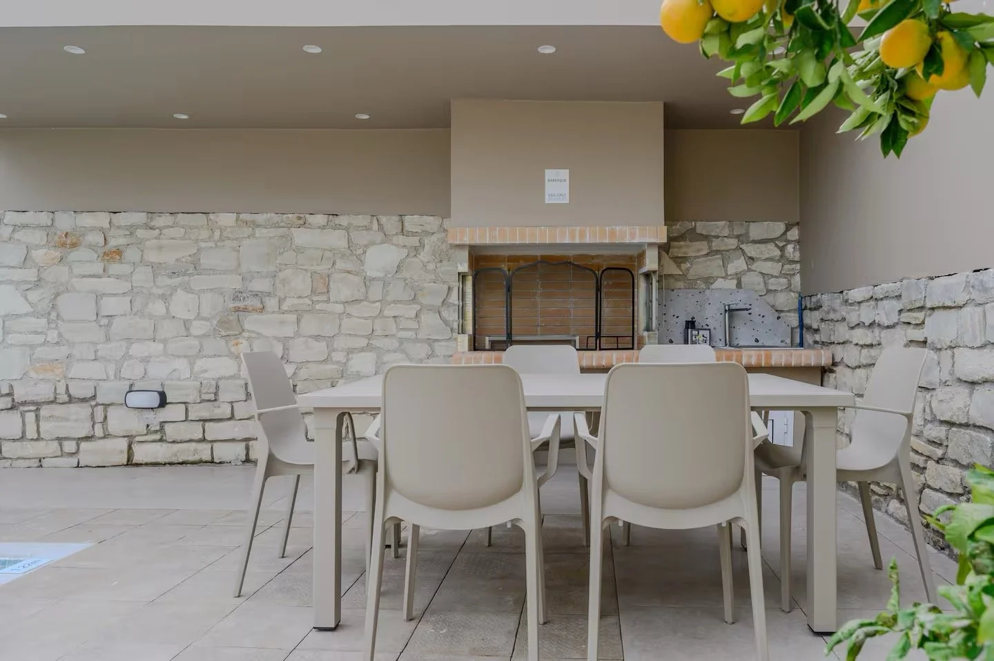 Outdoor dining area with a beige table and six matching chairs, stone wall, built-in brick fireplace, and grill, under a ceiling with recessed lighting, decorated with a lemon tree branch in the foreground.