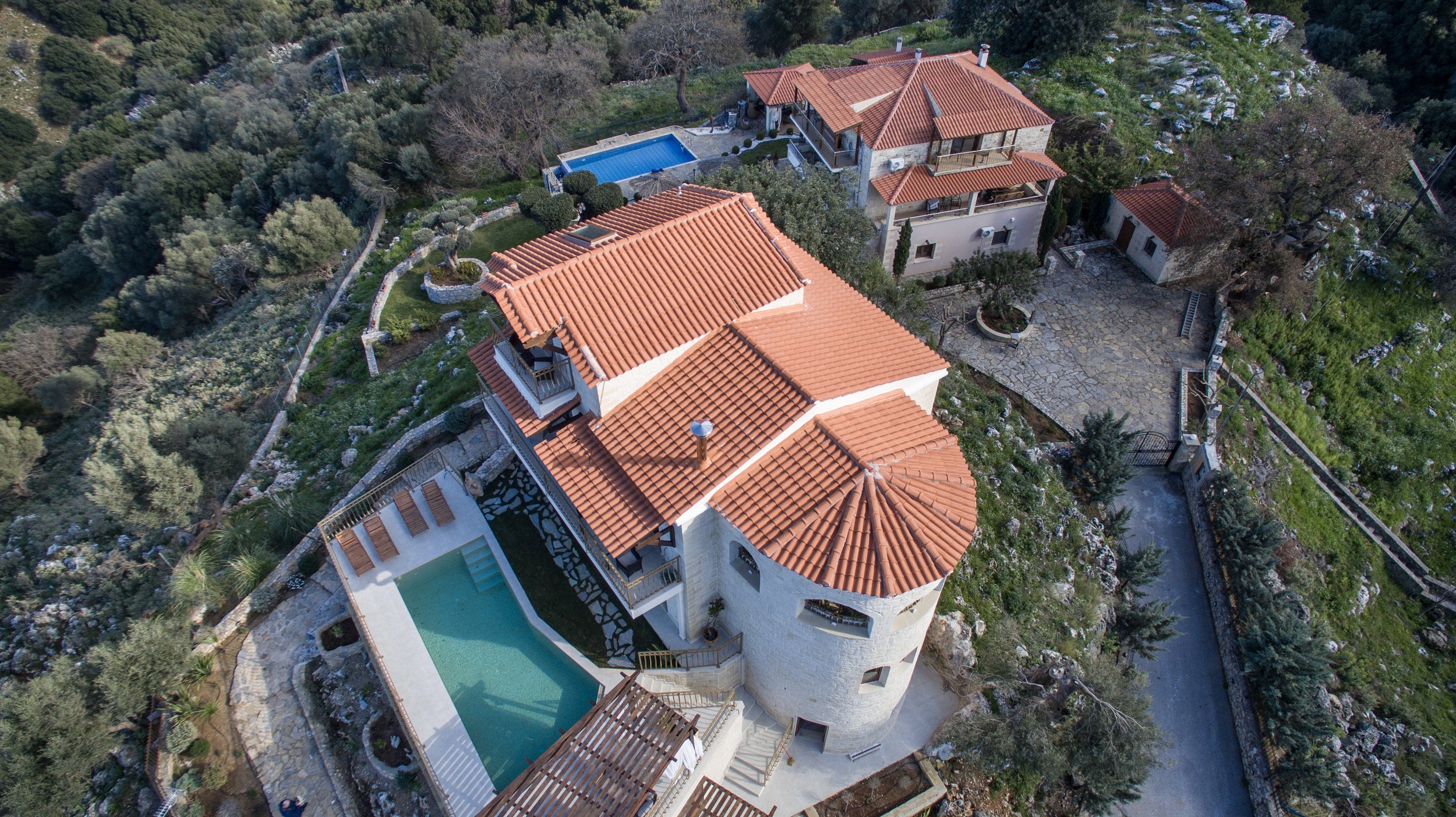Aerial view of a large property with red-tiled roofs, multiple buildings, a swimming pool, and surrounding greenery.