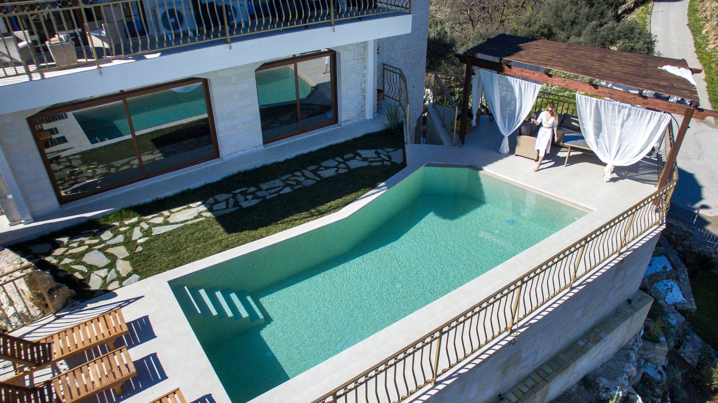 Aerial view of a modern villa with a swimming pool, wooden deck chairs, and a pergola-covered seating area.