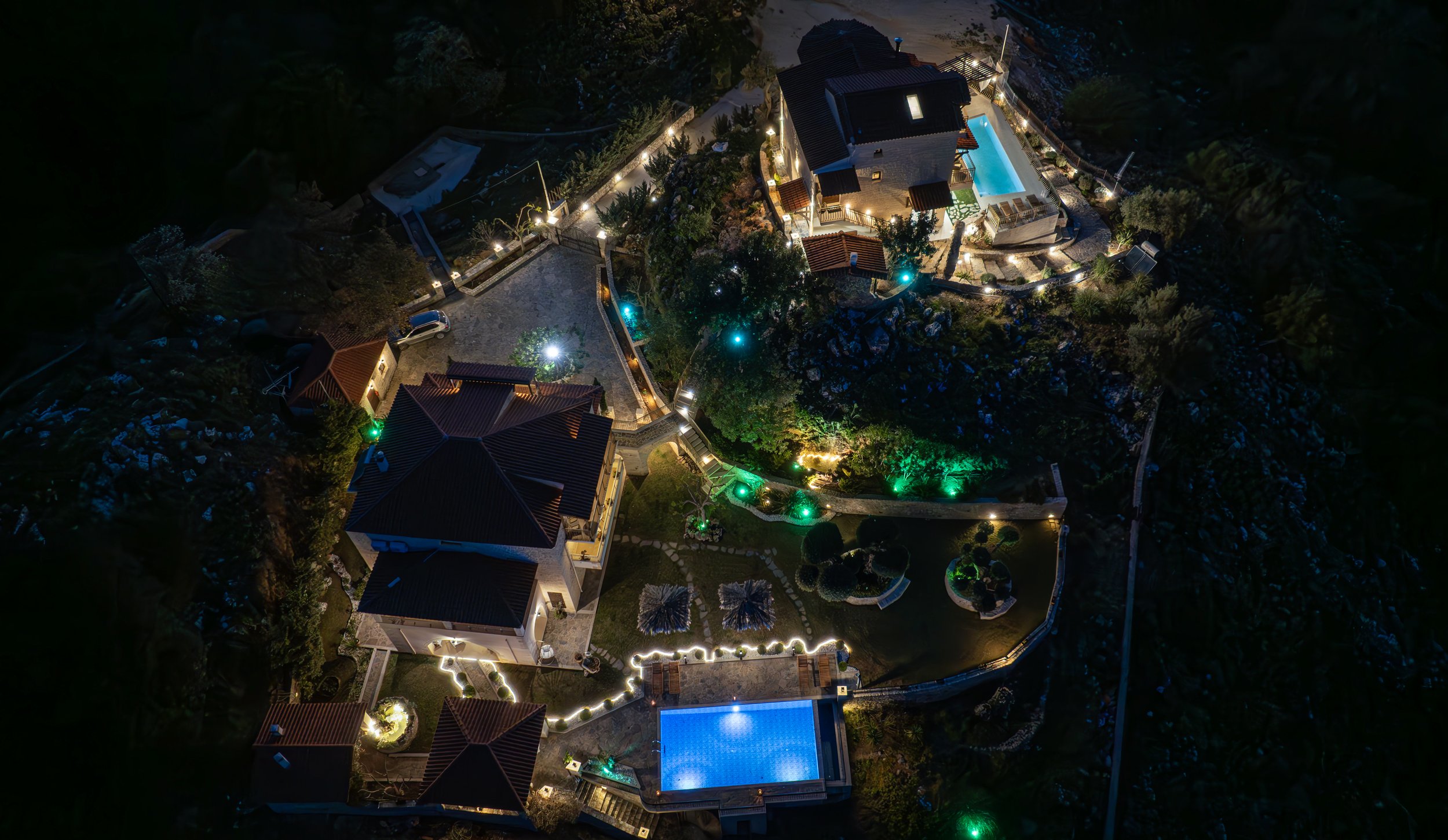 Aerial view of a luxurious villa complex at night, featuring multiple buildings and illuminated outdoor areas. Includes well-lit footpaths, landscaped gardens, and two swimming pools with blue lighting. Surrounded by natural scenery.