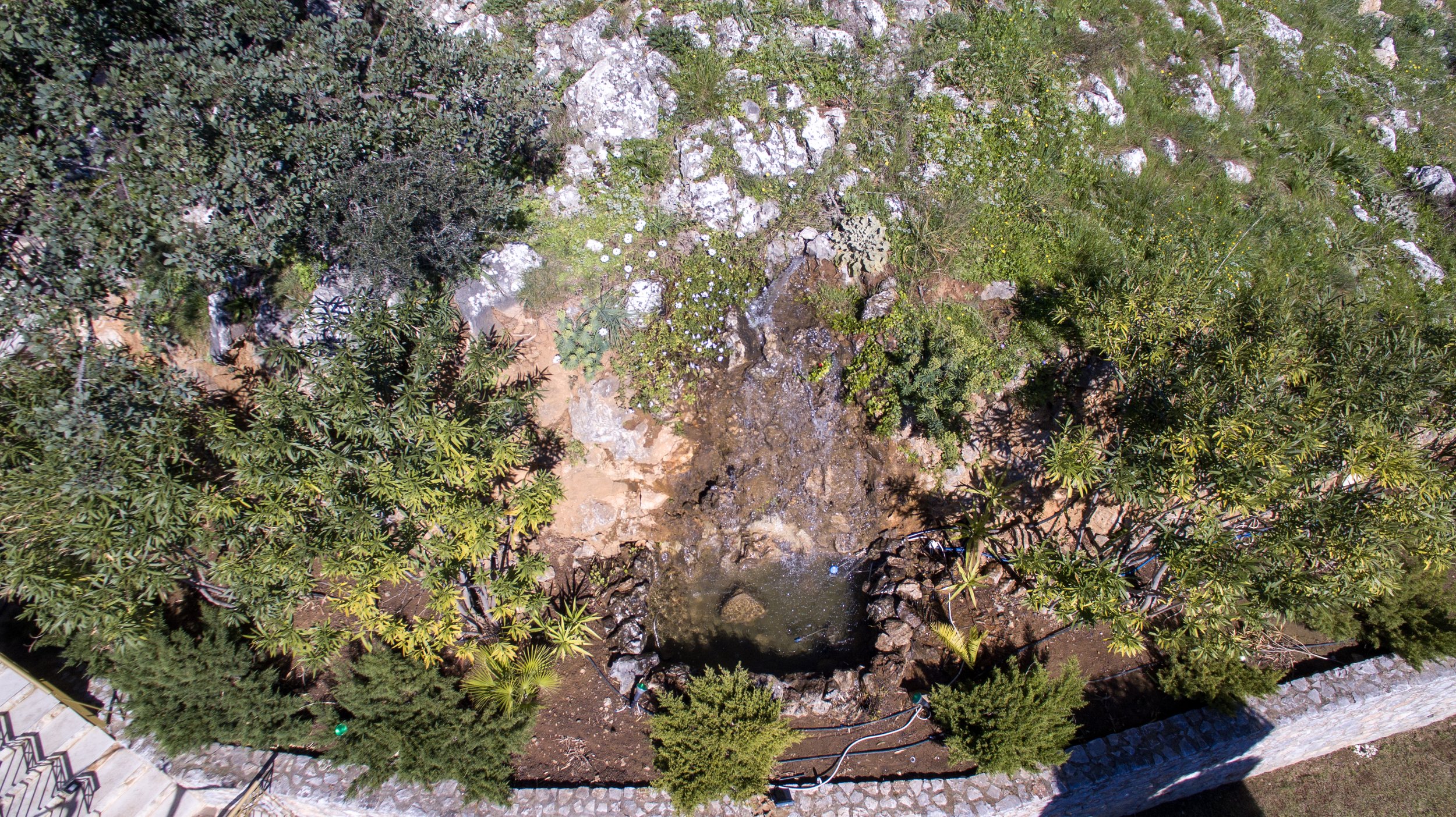 Aerial view of a small rocky waterfall surrounded by green foliage and trees in a garden setting. Water flows into a small pond bordered with rocks. Stone steps and a pathway are visible.