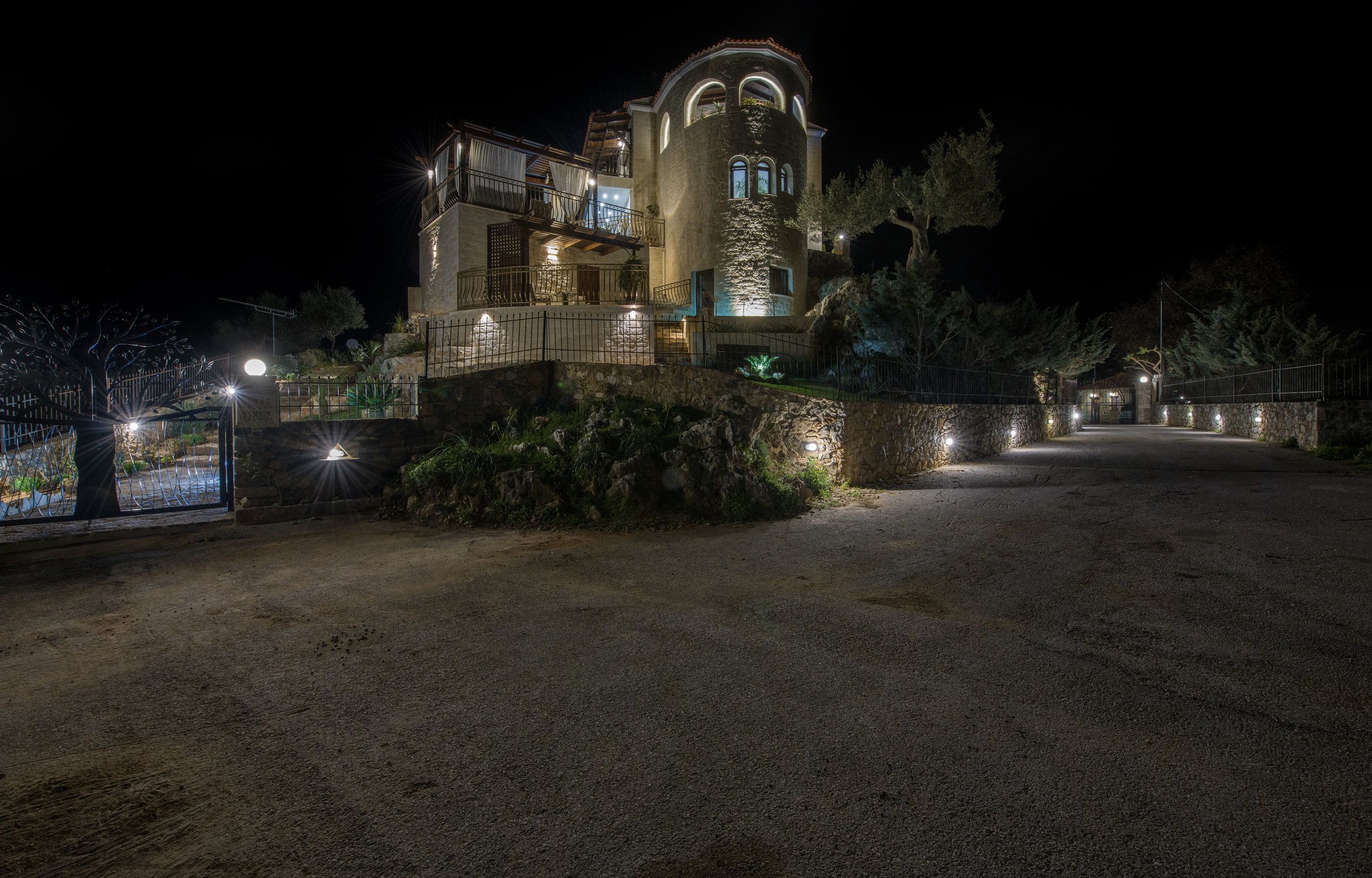 Illuminated stone house at night with surrounding garden and stone fence, lit by outdoor lights.