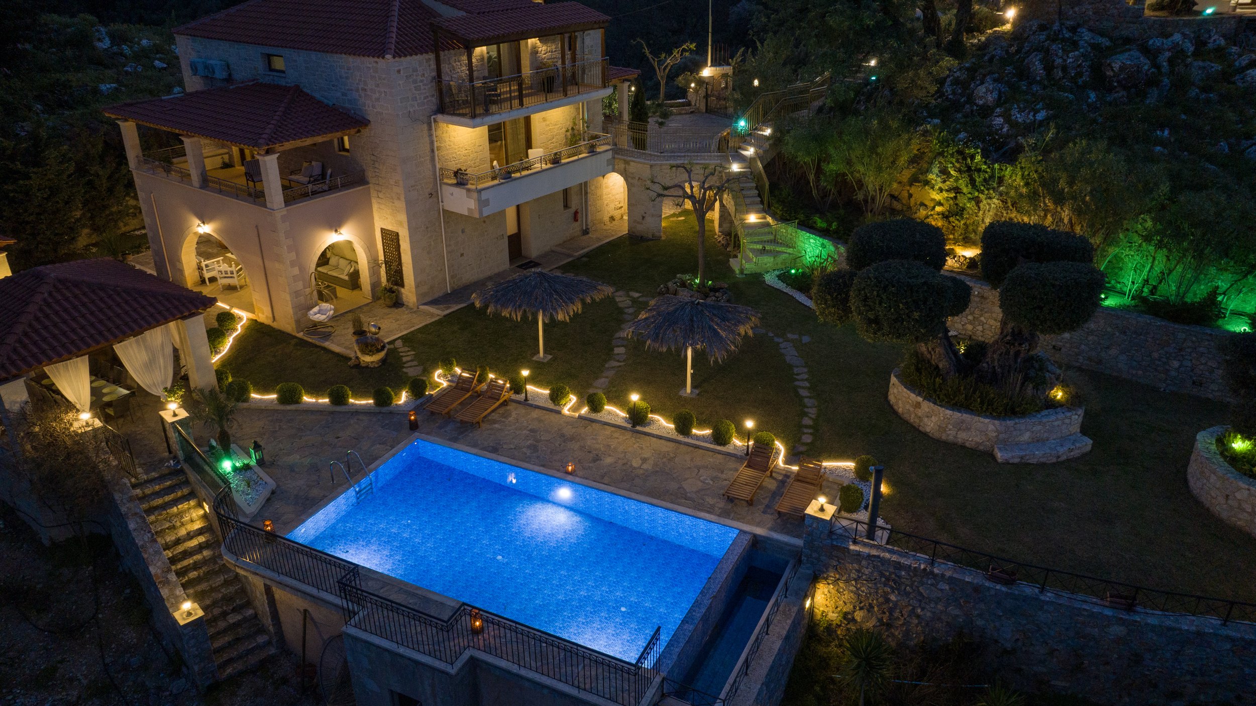 Aerial night view of a luxurious villa with a swimming pool, outdoor seating, and decorative lighting. The property features stone architecture and landscaped gardens with trees and pathways.
