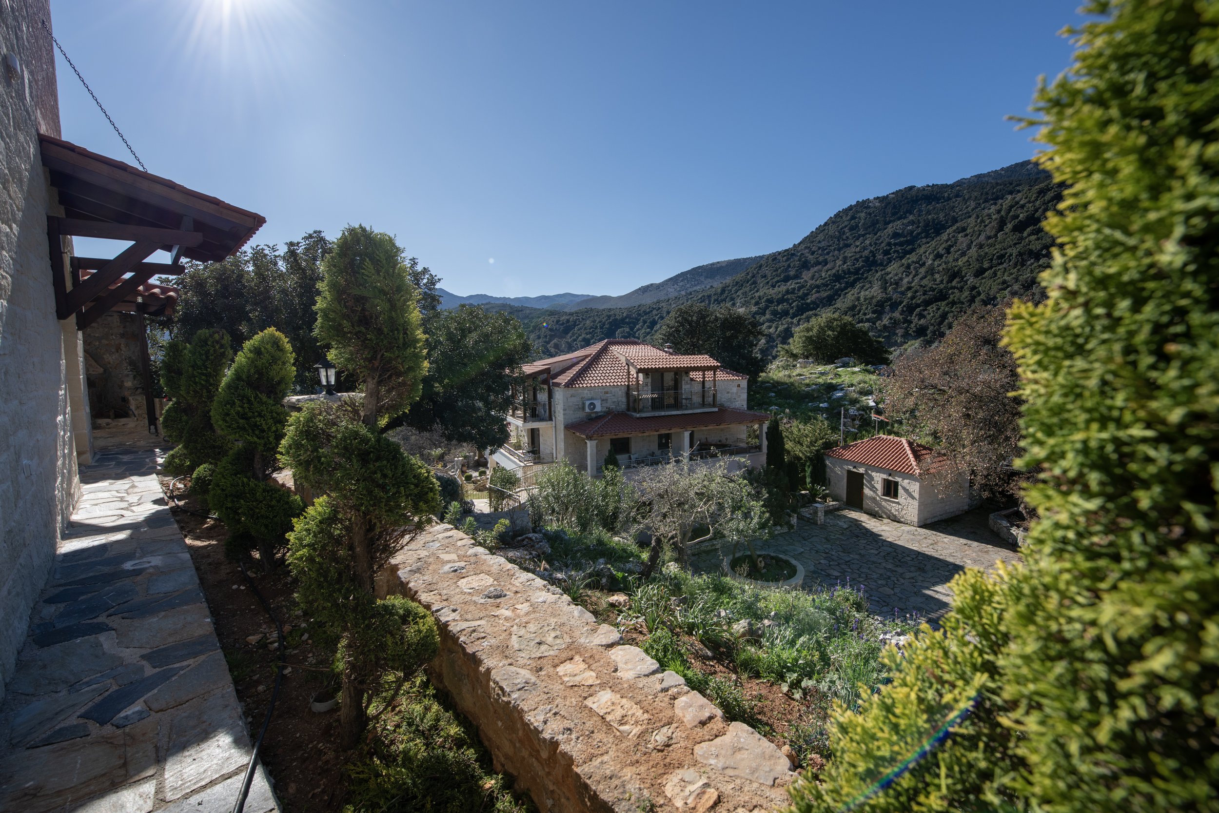 Stone house with red-tiled roof in a mountainous landscape, surrounded by trees and a garden, under a clear blue sky.