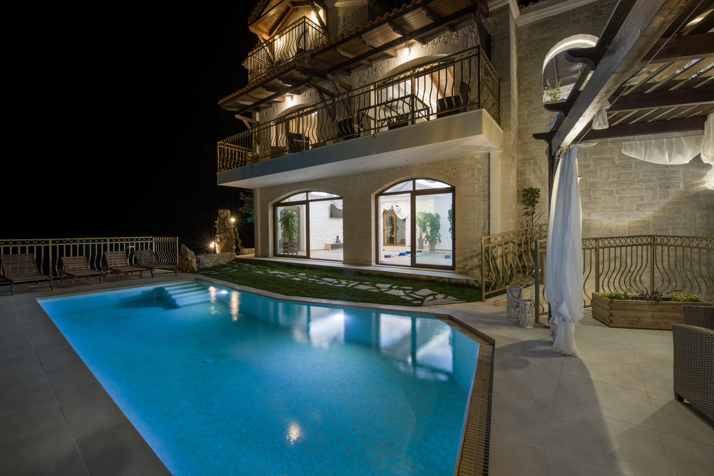 Luxury house with illuminated pool at night, featuring balconies, large windows, and outdoor seating.