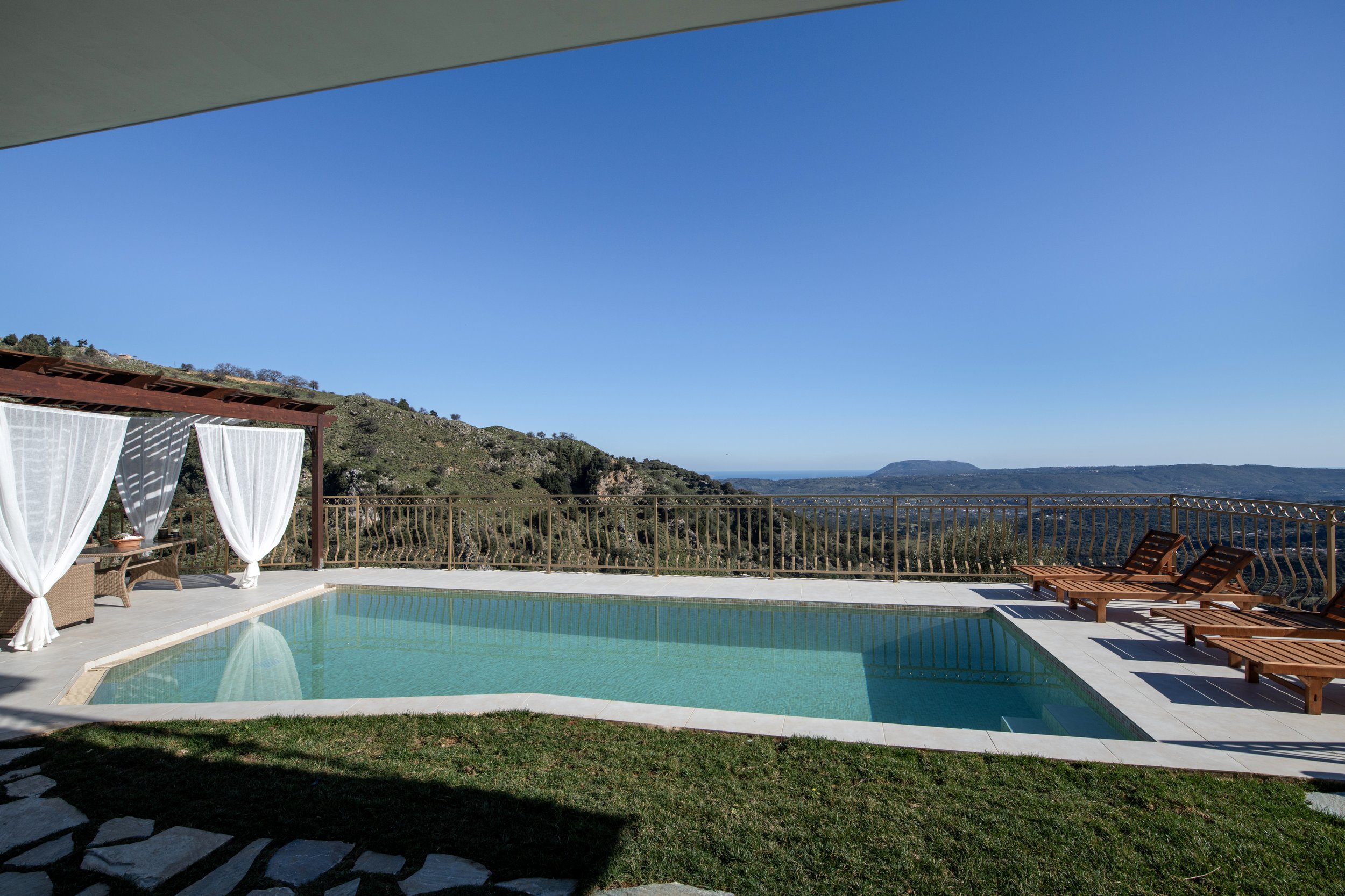 Outdoor pool with lounge chairs and a pergola with curtains, set in a landscape with mountains and clear sky.