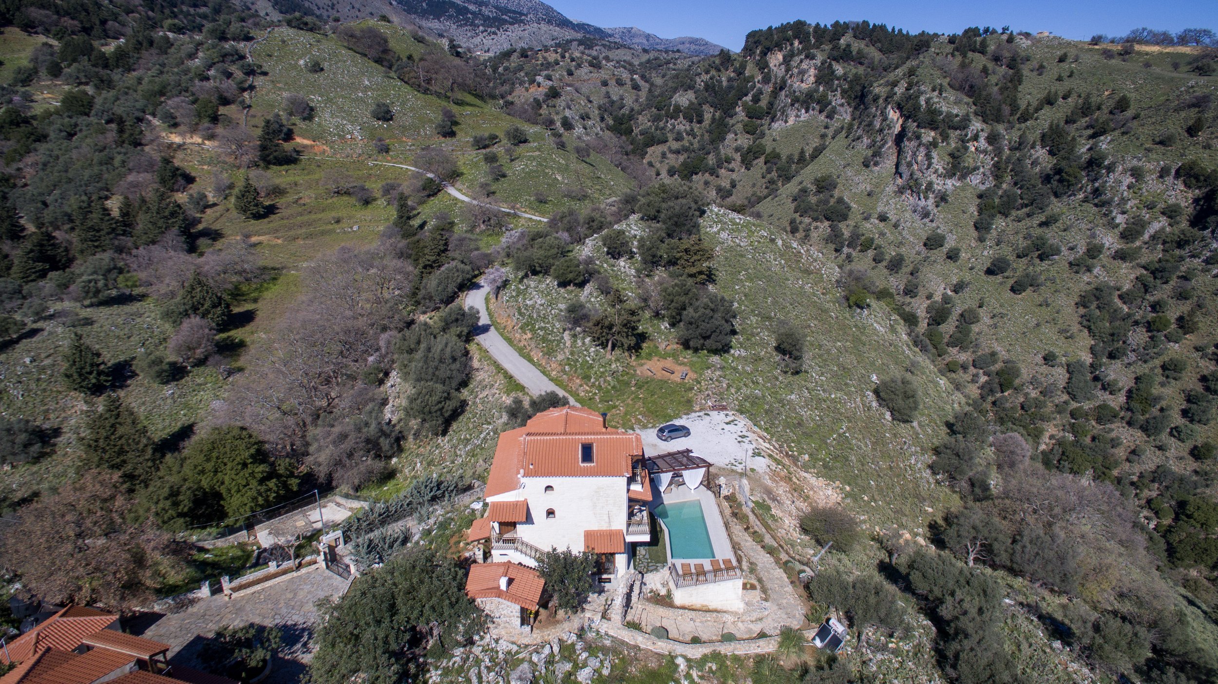 Aerial view of a villa with a terracotta roof, swimming pool, and surrounding greenery in a mountainous area.