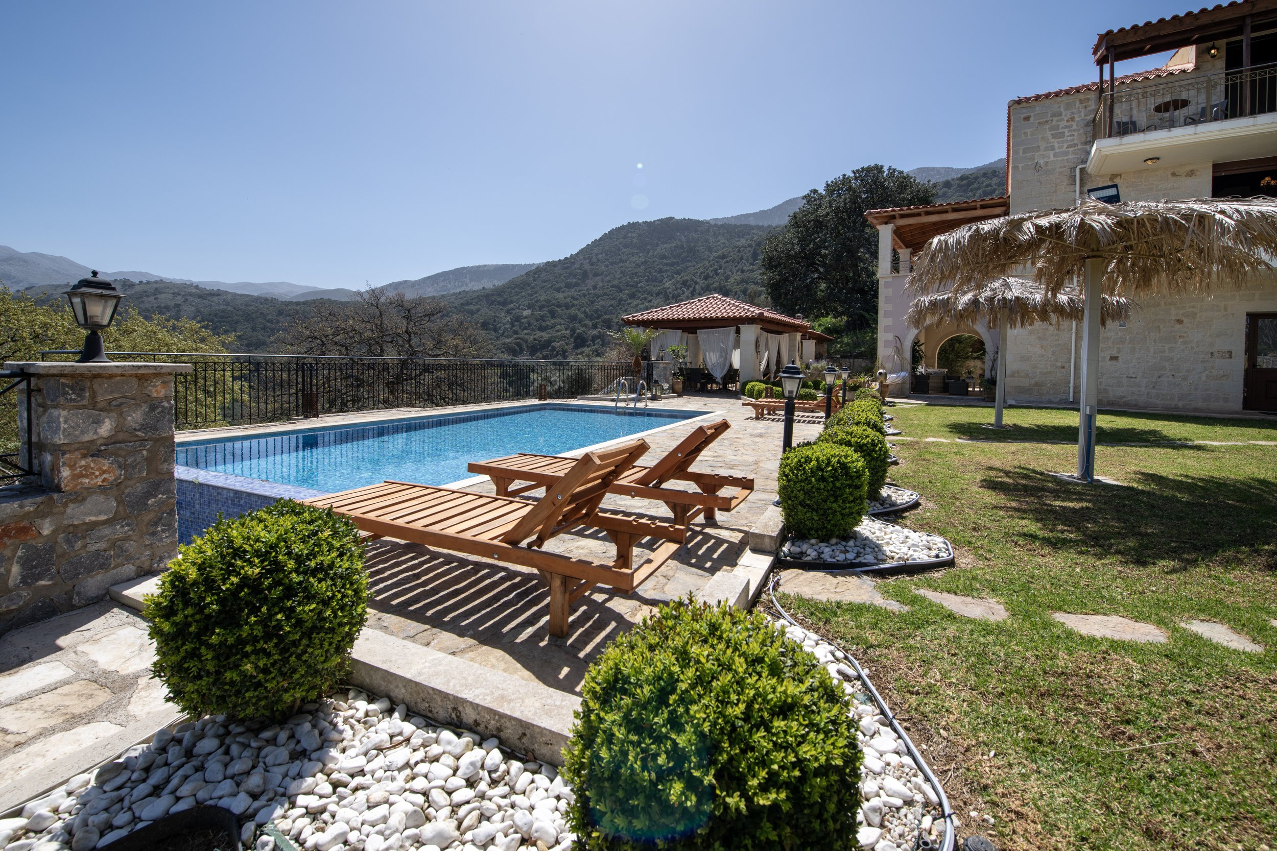 Outdoor pool area with wooden loungers, stone patio, decorative shrubs, and a gazebo; a house and mountains in the background.